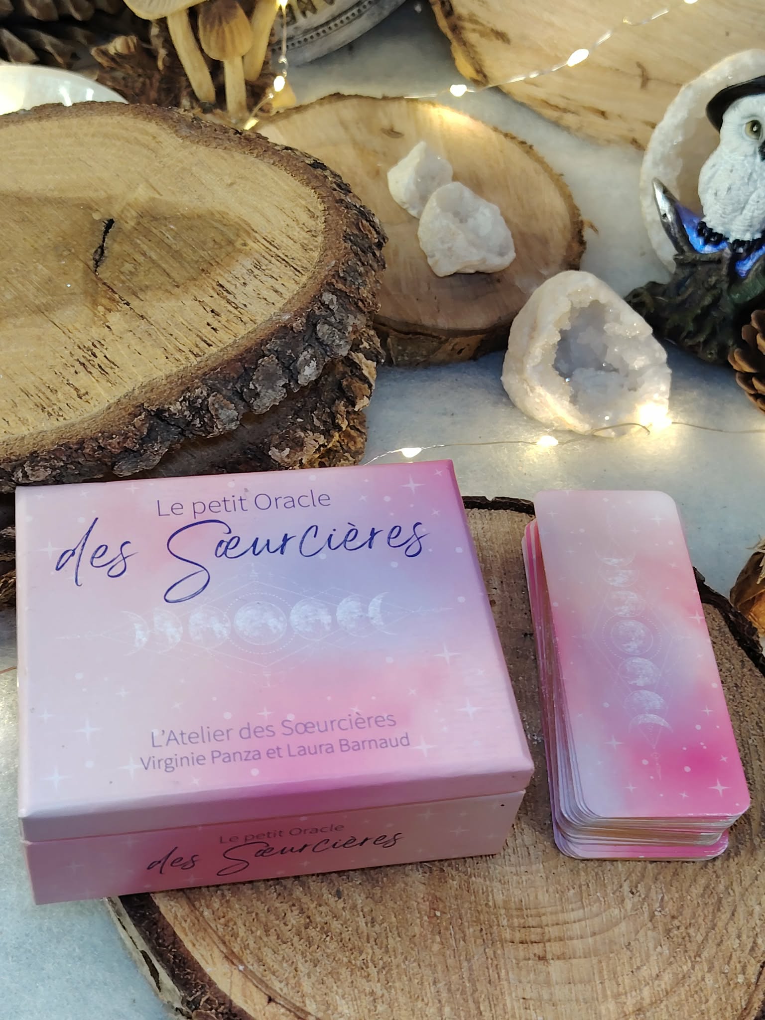 Le Petit Oracle des Sœurcières (L’Atelier des Sœurcières) : boîte rose fermée et paquet de cartes assorti posés sur rondin de bois, décor avec pierres de quartz blanc et ambiance cocooning