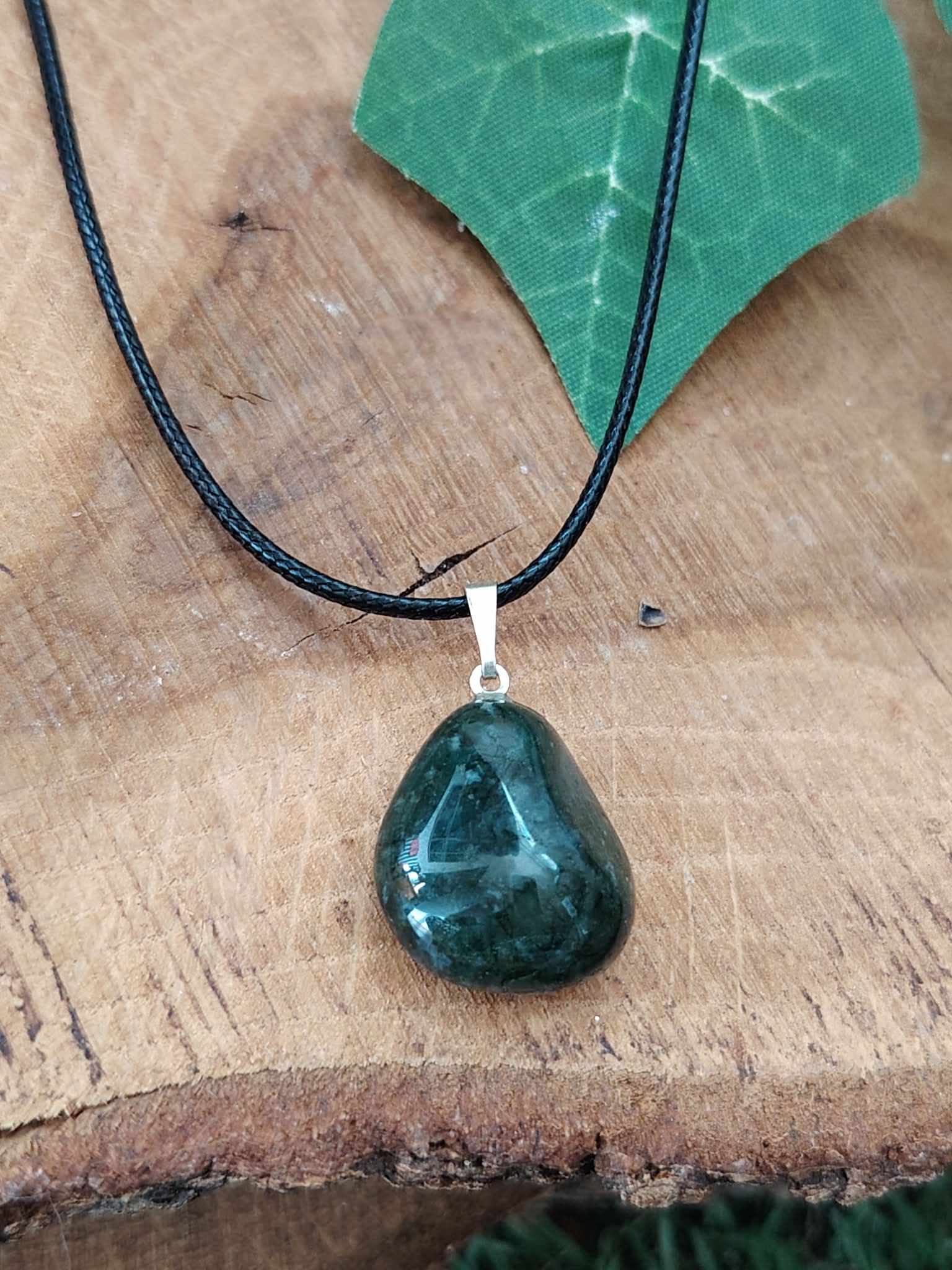 Pendentif agate mousse vert foncé sur cordon noir, posé sur tranche de bois avec feuille verte, sérénité affective et stabilité du cœur