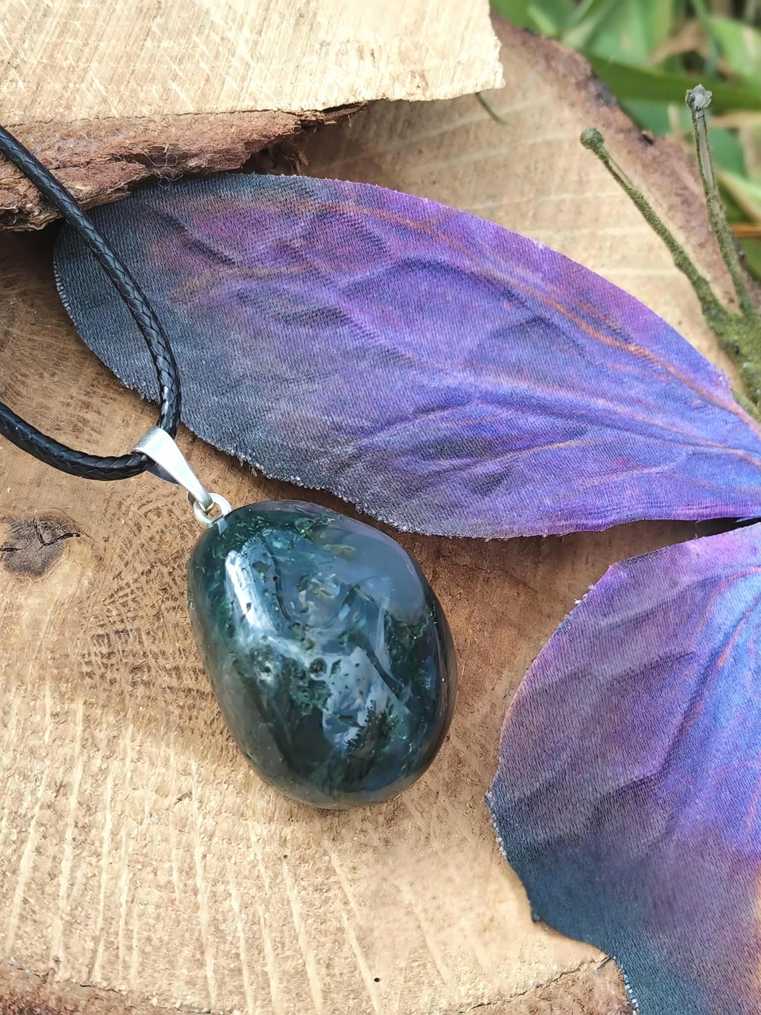 Pendentif agate mousse vert-bleuté sur cordon noir, photographié sur tranche de bois avec grande feuille violette, douceur du cœur et sérénité intérieure