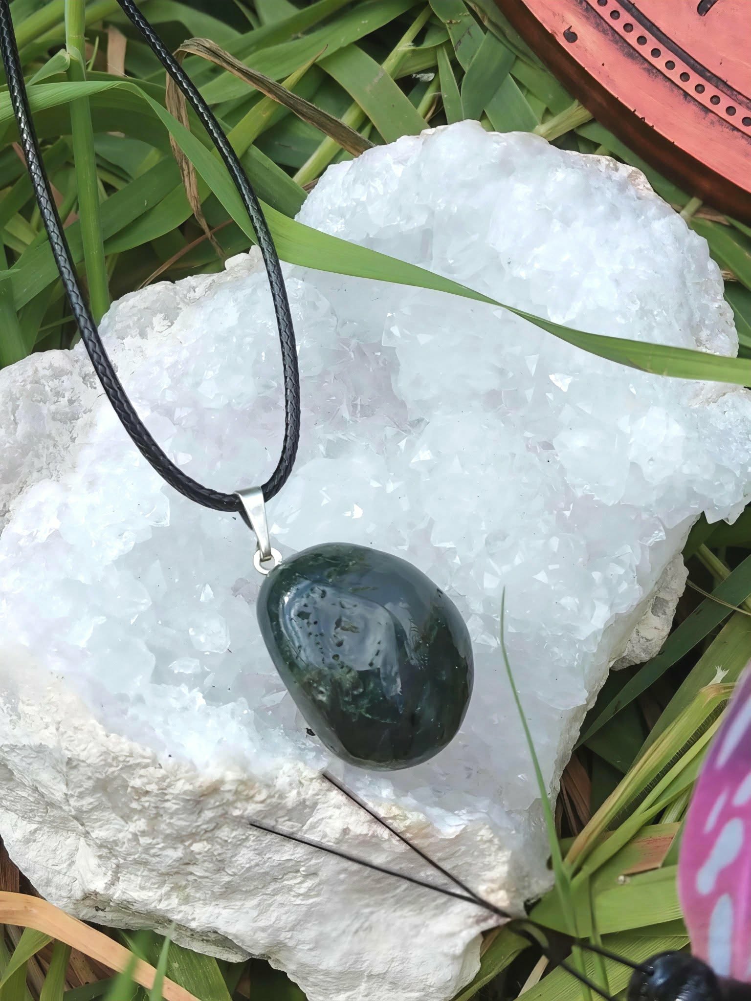 Pendentif agate mousse vert foncé sur cordon noir, posé sur galet blanc avec herbe et touche violette, recentrage doux et paix émotionnelle