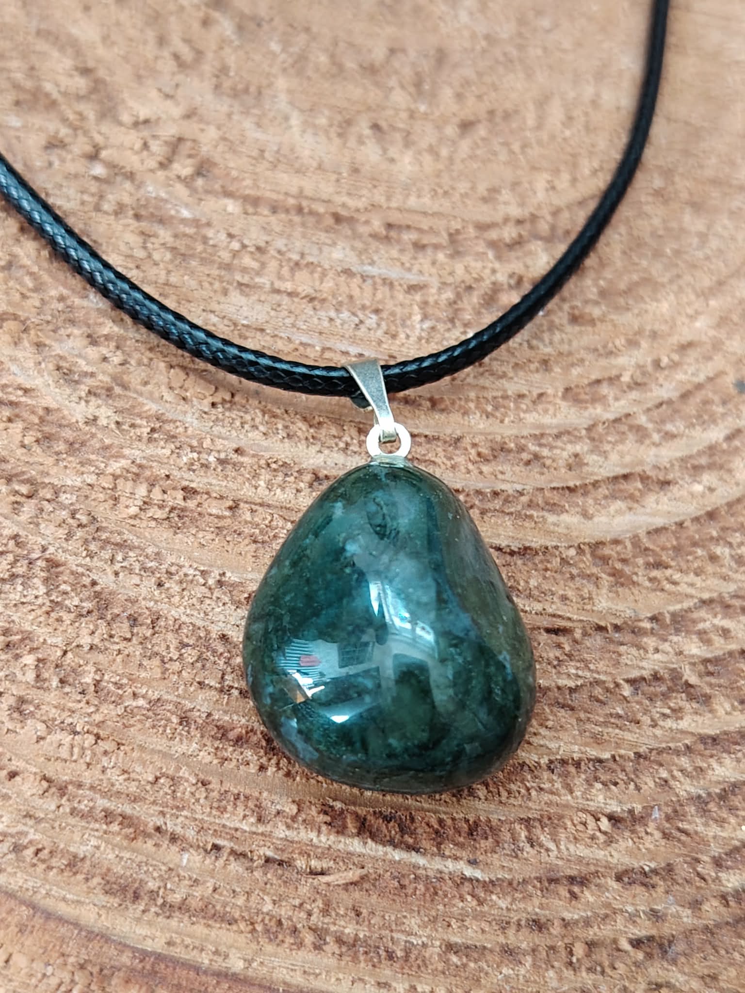 Gros plan sur pendentif agate mousse vert mousse sur cordon noir, posé sur rondin de bois, clarté du ressenti et sérénité douce