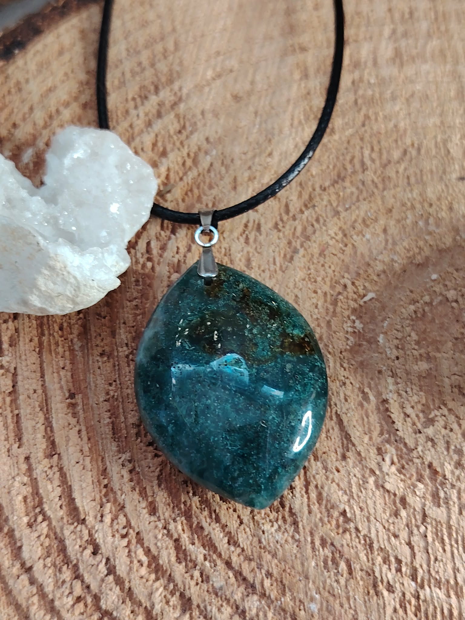 Pendentif en agate mousse poli (forme ronde) sur cordon noir, posé sur un fond en bois avec une petite géode à côté ; pierre associée au chakra cœur, réputée pour apporter apaisement, harmonie émotionnelle et connexion à la nature.