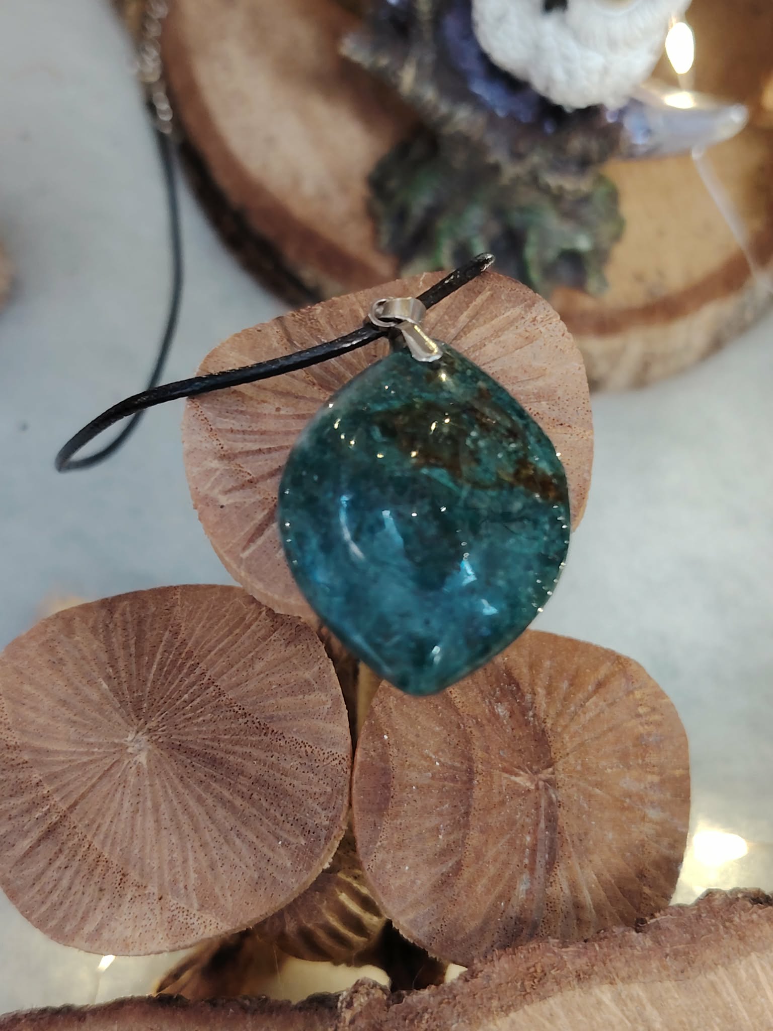 Pendentif agate mousse (goutte) sur cordon noir, posé sur galets ronds avec feuillage en arrière-plan ; pierre du chakra cœur, invite au calme et à l’équilibre émotionnel.