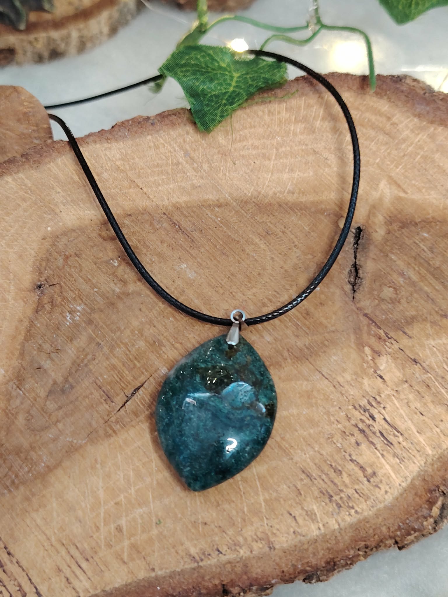 Pendentif agate mousse (goutte) sur cordon noir, posé sur tranche de bois avec feuille verte ; aide à se recentrer, apaiser et renforcer le lien à la nature.
