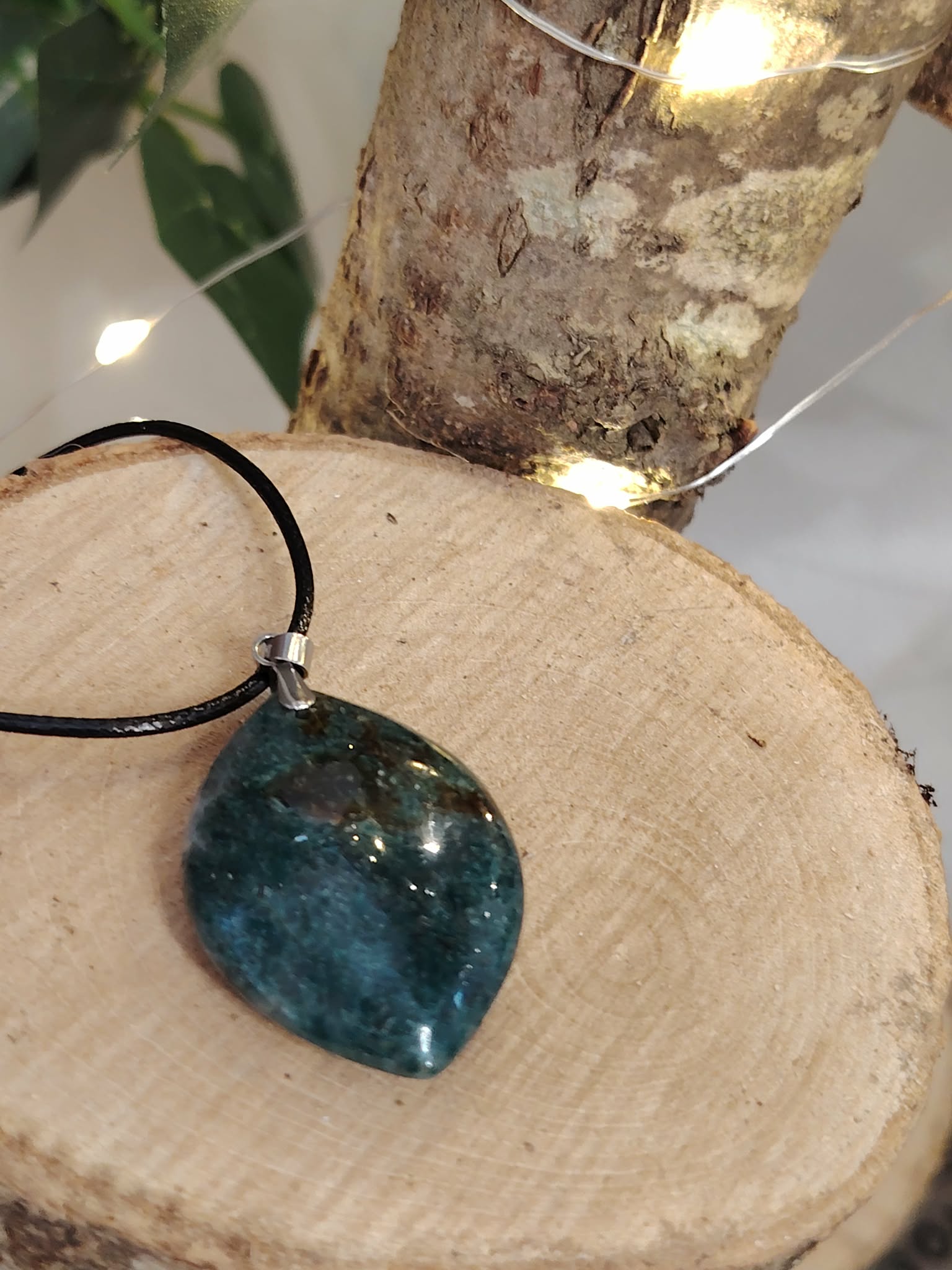 Pendentif agate mousse (goutte) sur cordon noir, posé sur support en bois avec feuillage ; favorise apaisement et ancrage naturel.