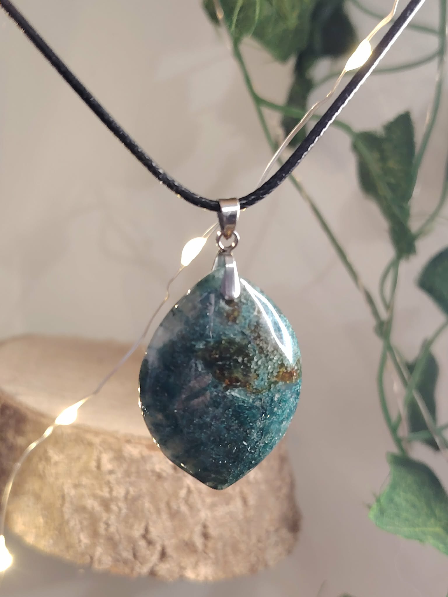 Pendentif en agate mousse (goutte) sur cordon noir, sur fond clair avec feuillage et support bois ; aide à l’apaisement et à la connexion à la nature