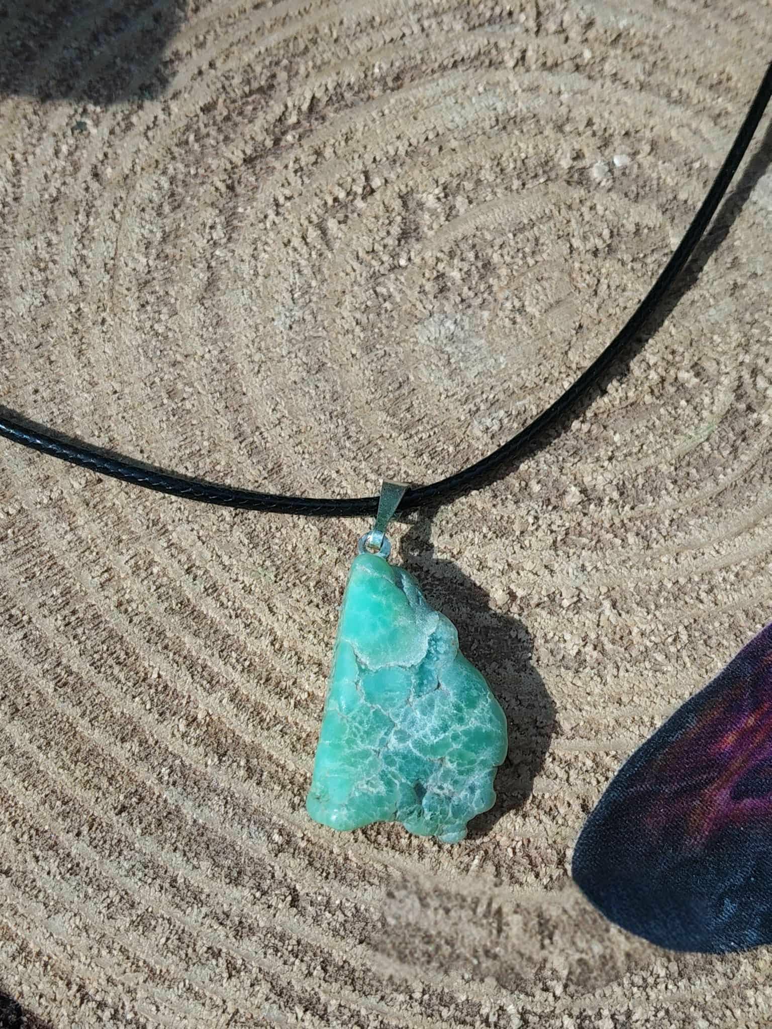 Pendentif chrysoprase brute vert d’eau sur cordon noir, posé sur tranche de bois aux cernes marqués, pierre naturelle associée au discernement, au calme des pensées et à la présence à soi.