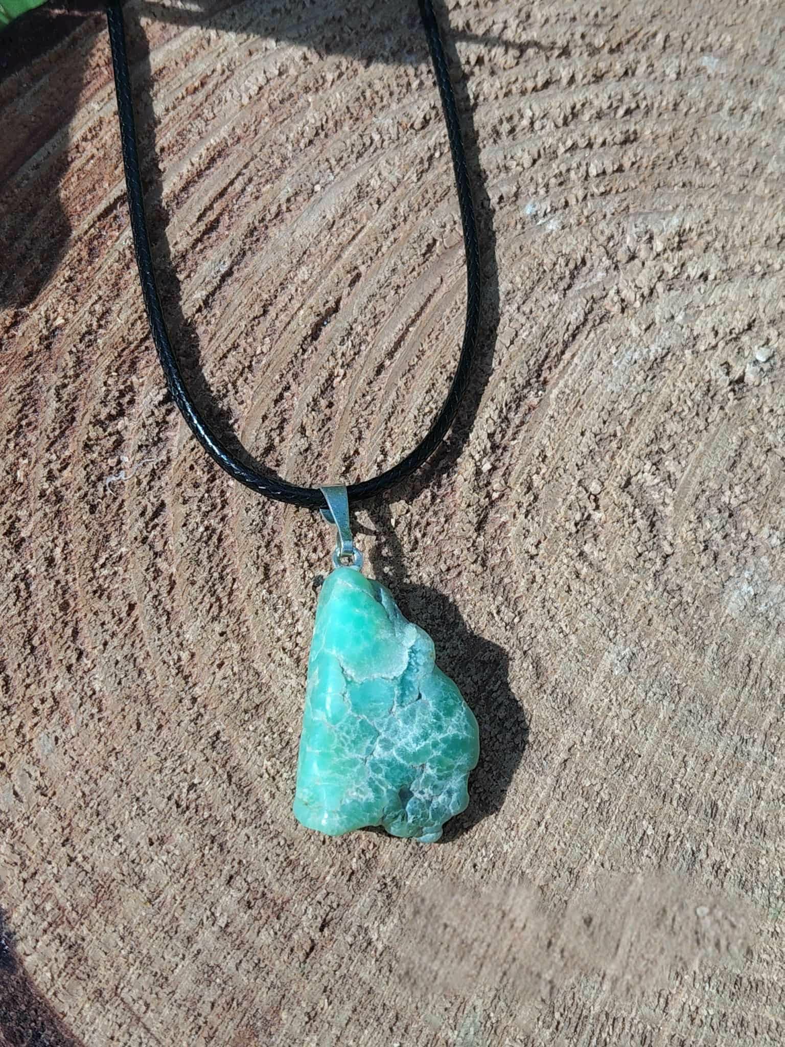 Pendentif chrysoprase naturelle brute sur cordon noir, posé sur bois aux veines marquées, bijou en pierre naturelle lié à la sensibilité apaisée, à l’harmonie du cœur et au réconfort intérieur.