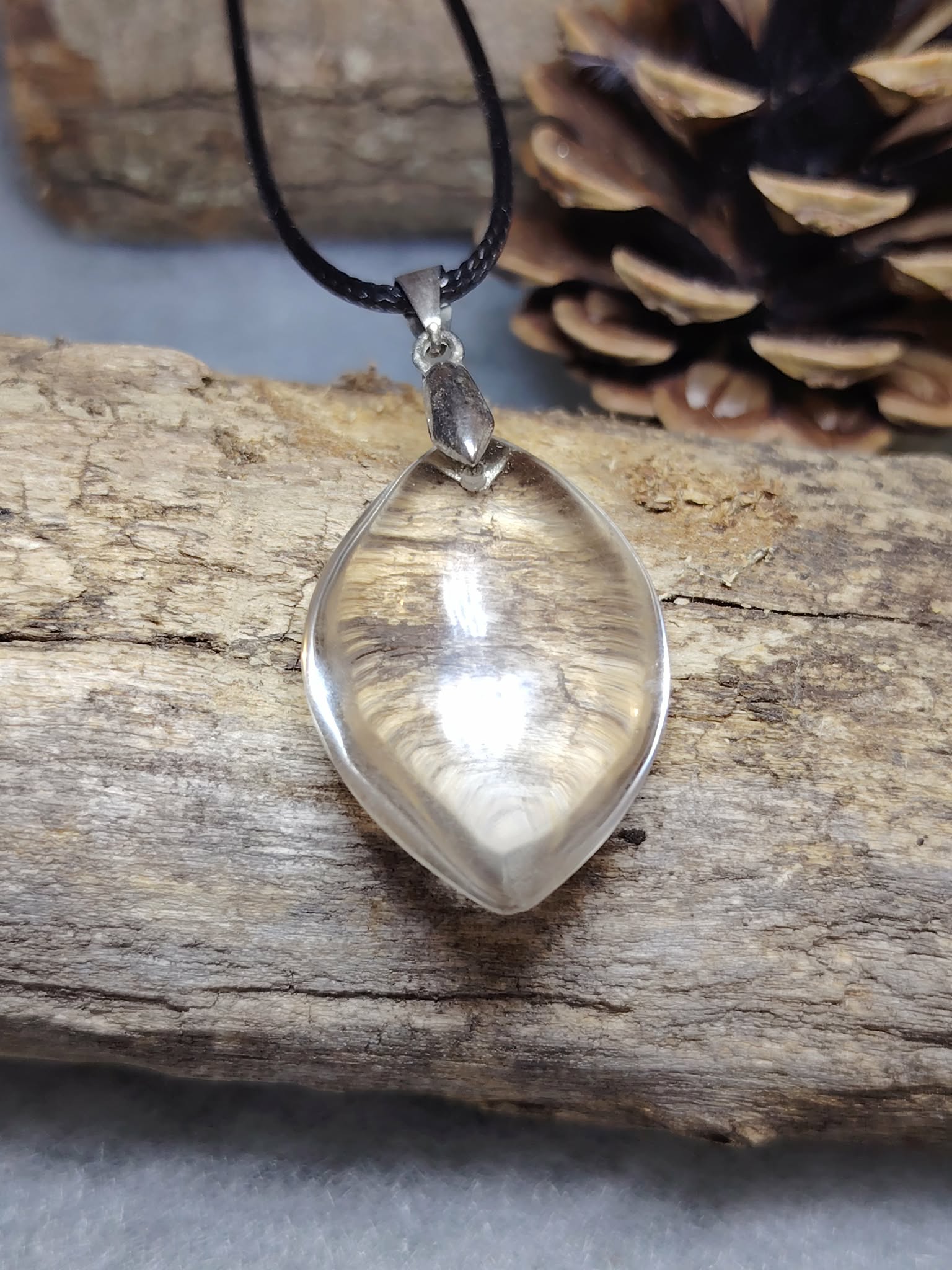 pendentif goutte cristal de roche quartz transparent sur cordon noir posé sur bois brut avec pomme de pin en arrière-plan — clarté d’esprit