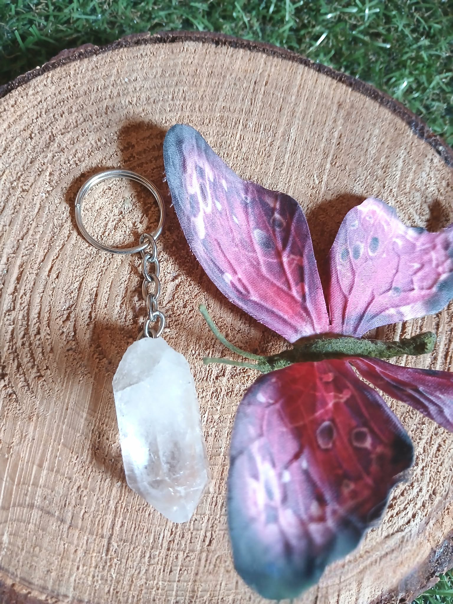 Porte-clés / bijou de sac en cristal de roche taillé en pointe facettée, avec anneau métal, posé sur une tranche de bois avec un papillon décoratif violet et rose, ambiance nature