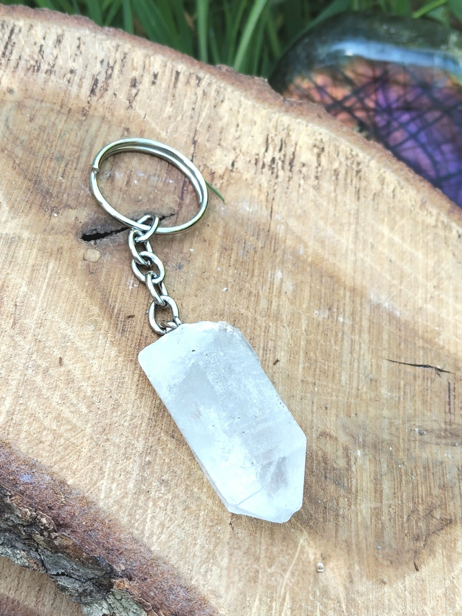 Porte-clés / bijou de sac en cristal de roche taillé en pointe facettée, avec chaîne et anneau métal, posé sur une tranche de bois clair en décor naturel