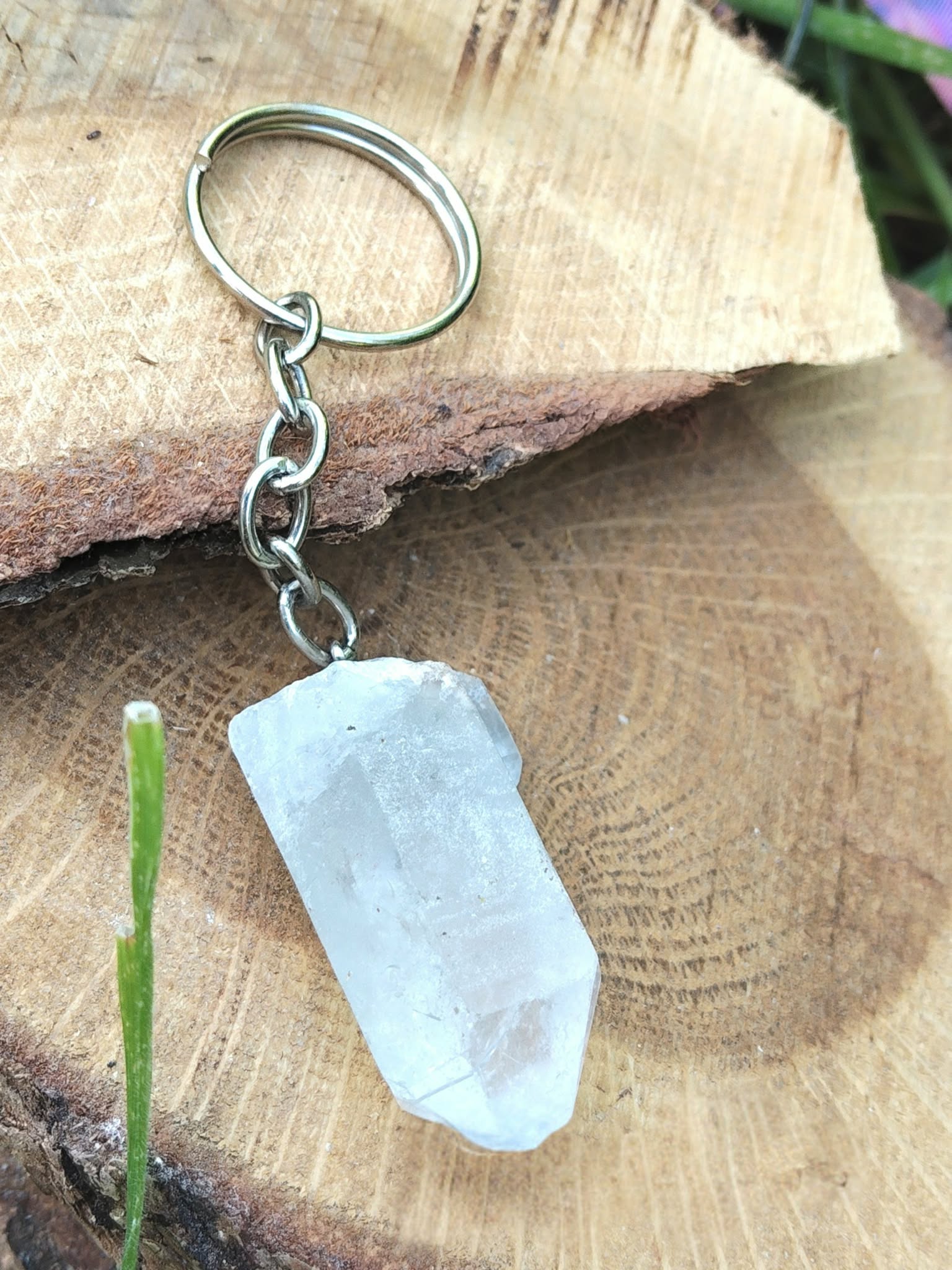 Porte-clés / bijou de sac en cristal de roche taillé en pointe facettée, avec chaîne et anneau métal, suspendu devant une tranche de bois clair avec une petite tige verte en décor naturel