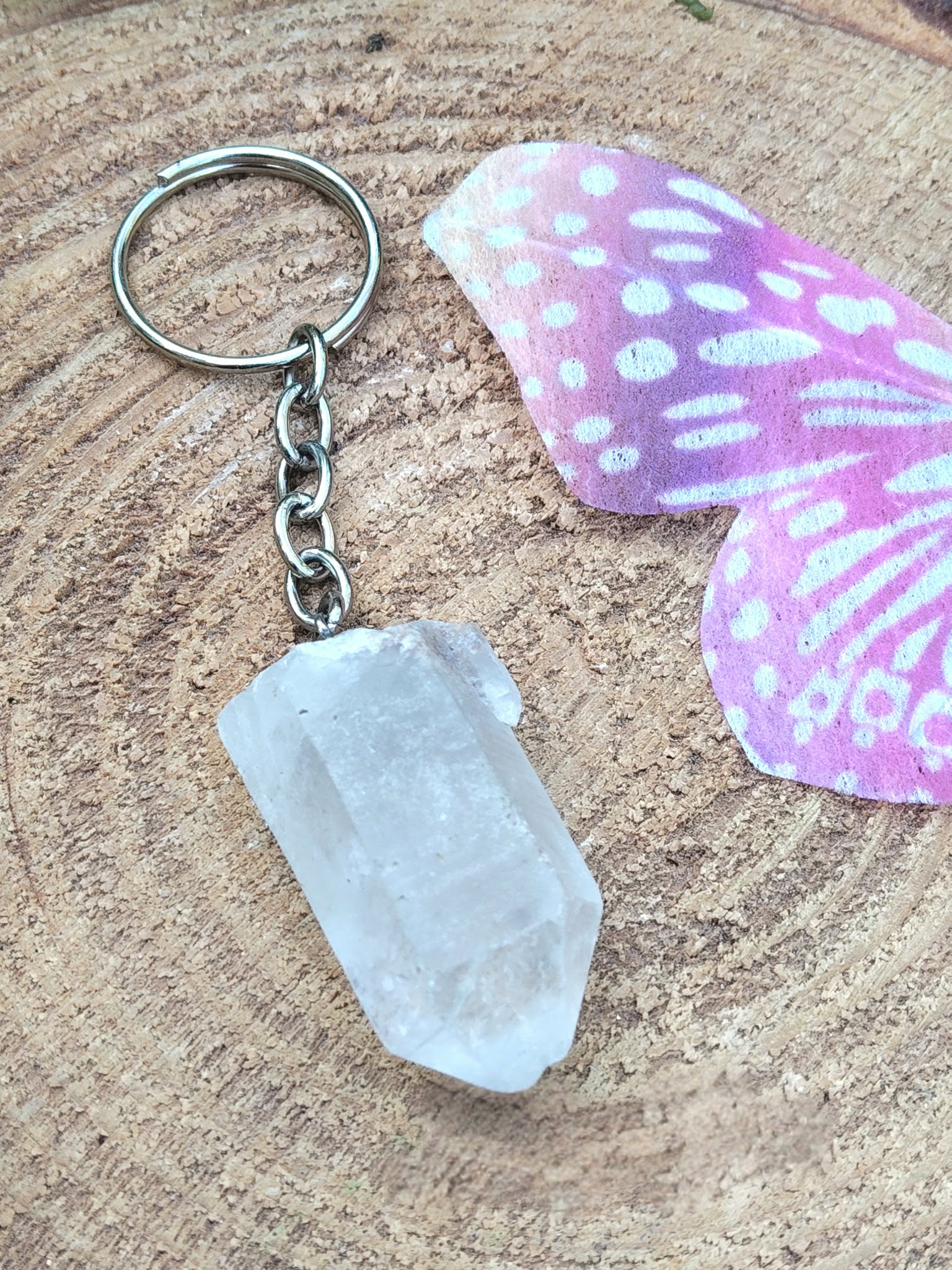 Porte-clés / bijou de sac en cristal de roche taillé en pointe facettée, avec anneau métal, posé sur une tranche de bois avec deux petits champignons décoratifs