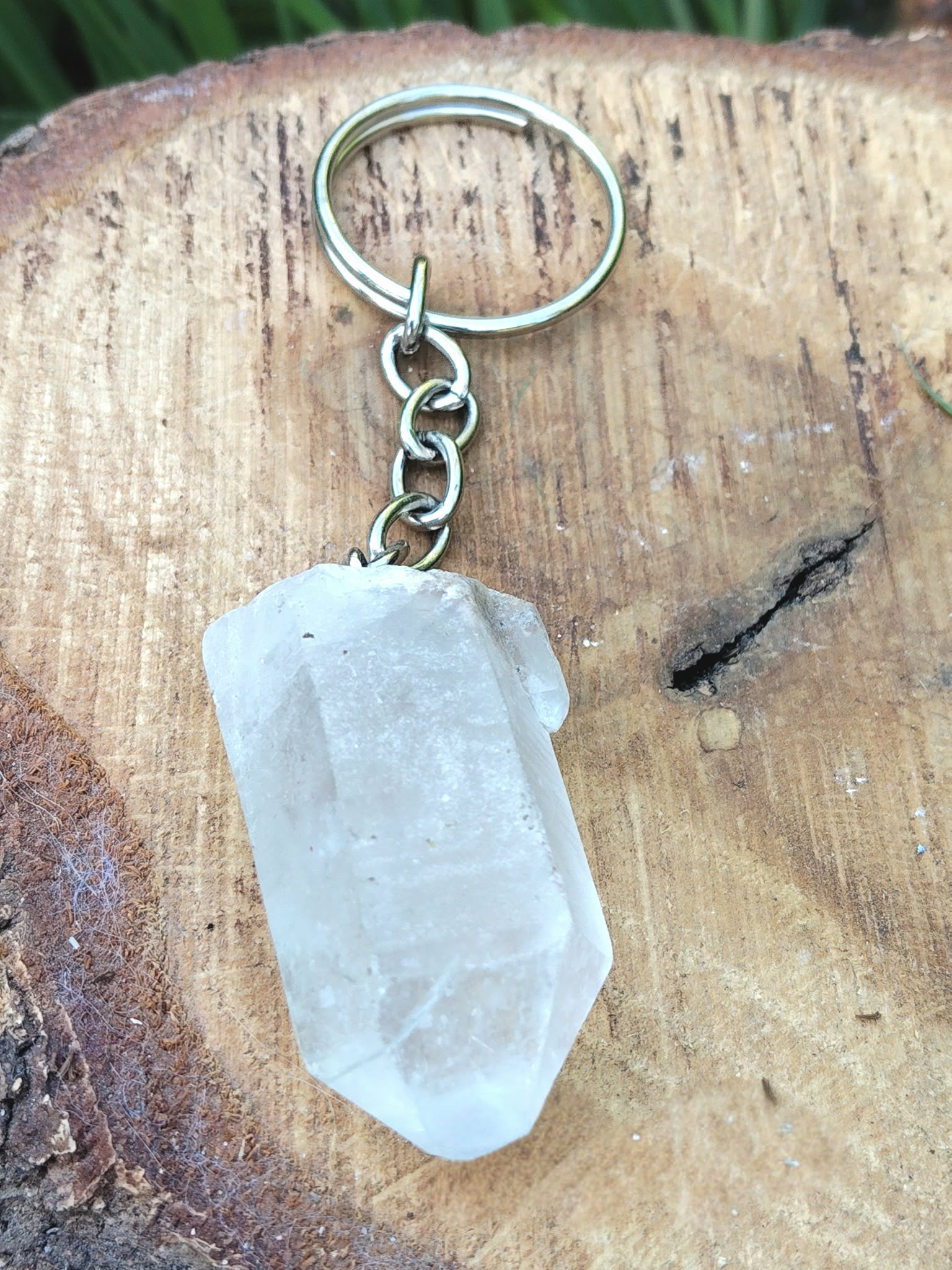 Porte-clés / bijou de sac en cristal de roche taillé en pointe facettée, avec anneau métallique, posé sur une tranche de bois clair en décor naturel