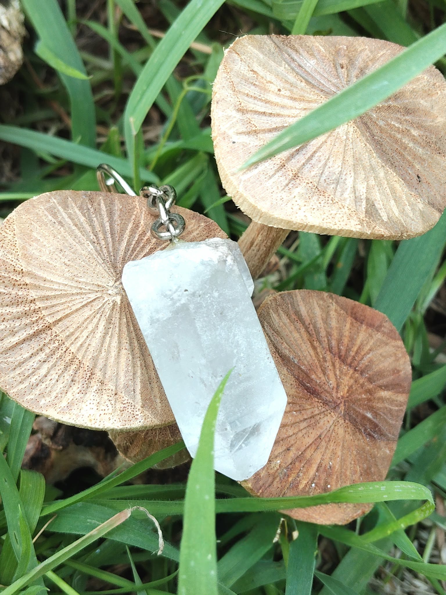 Porte-clés / bijou de sac en cristal de roche taillé en pointe facettée, posé sur des feuilles rondes beiges au milieu de brins d’herbe verte, décor nature