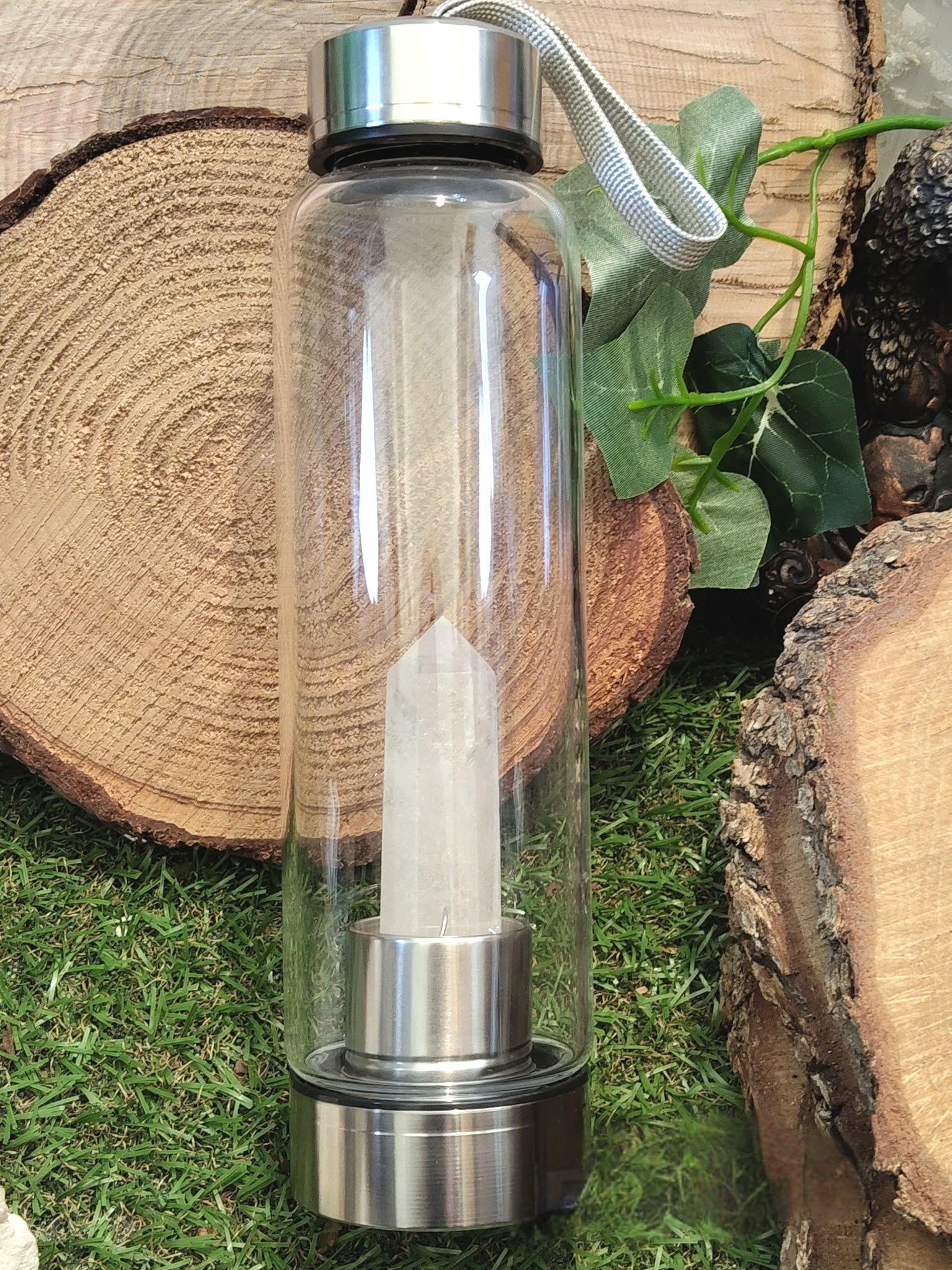 bouteille deau en verre énergisant cristal de roche, vue entière, bouchon inox avec dragonne, pointe de cristal de roche intégrée, décor rondins de bois et feuillage