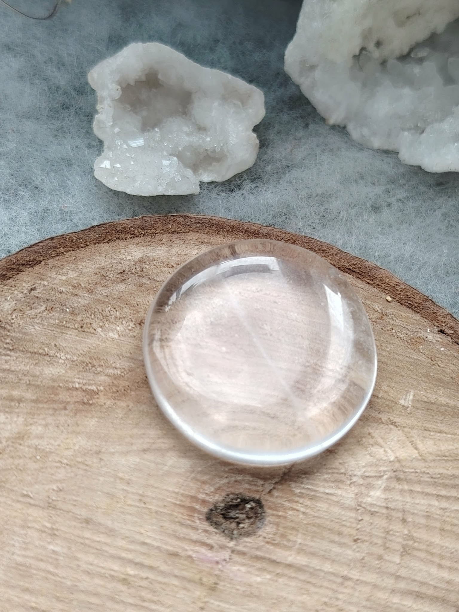Galet cristal de roche poli posé sur tranche de bois avec quartz brut en arrière-plan, pierre roulée translucide associée à la pureté, à l’amplification et à l’harmonie intérieure.