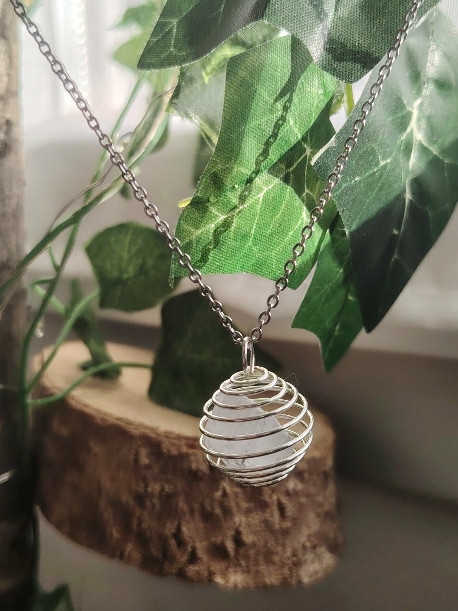 Pendentif cage spirale avec quartz blanc (cristal de roche), suspendu sur petit support en bois, décor plante verte, énergie de clarté.