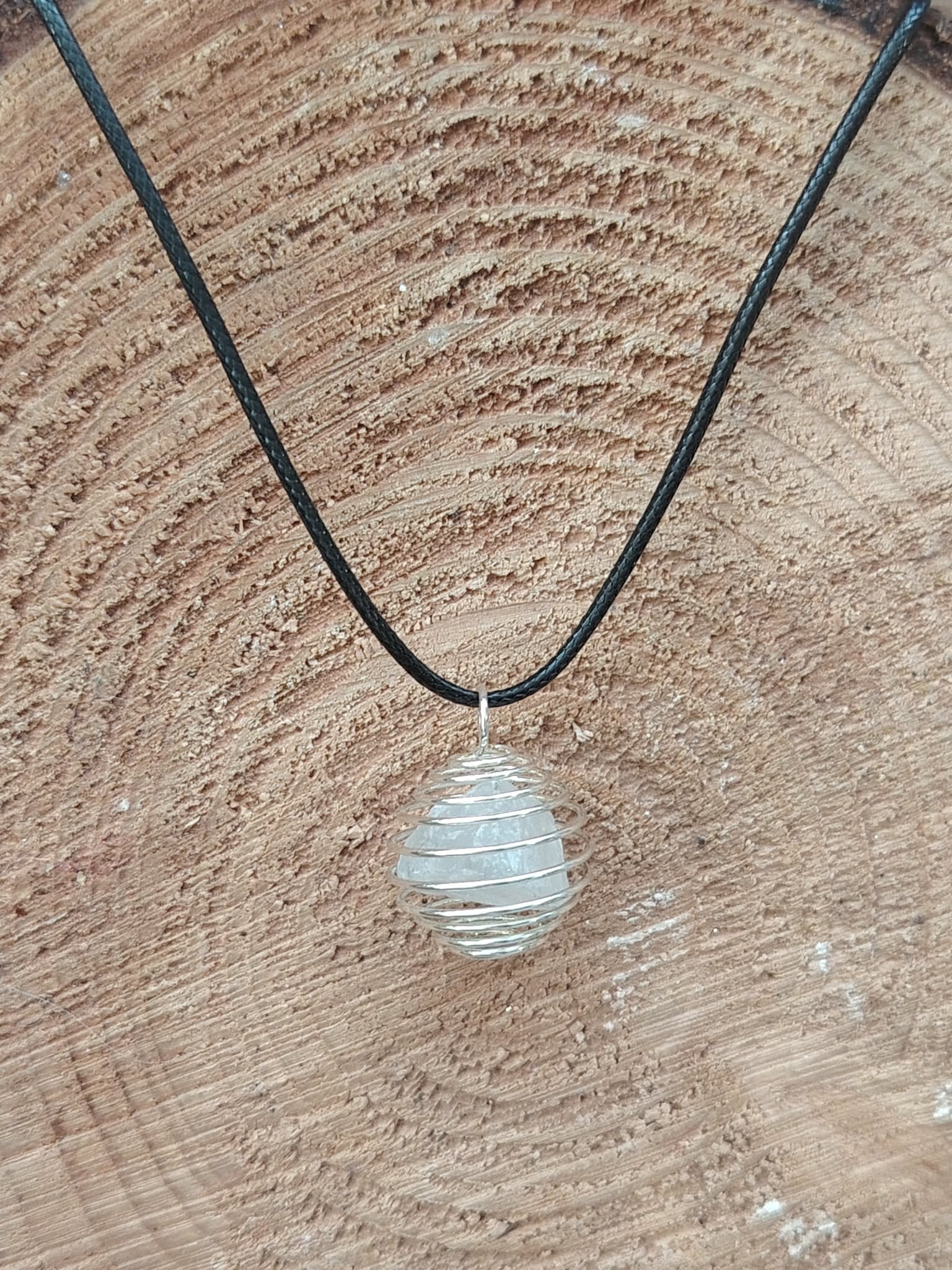 Pendentif cage spirale avec amas de quartz / cristal de roche, sur cordon noir, posé sur tranche de bois — associé à la clarté, l’harmonisation et l’amplification des intentions (symbolique).