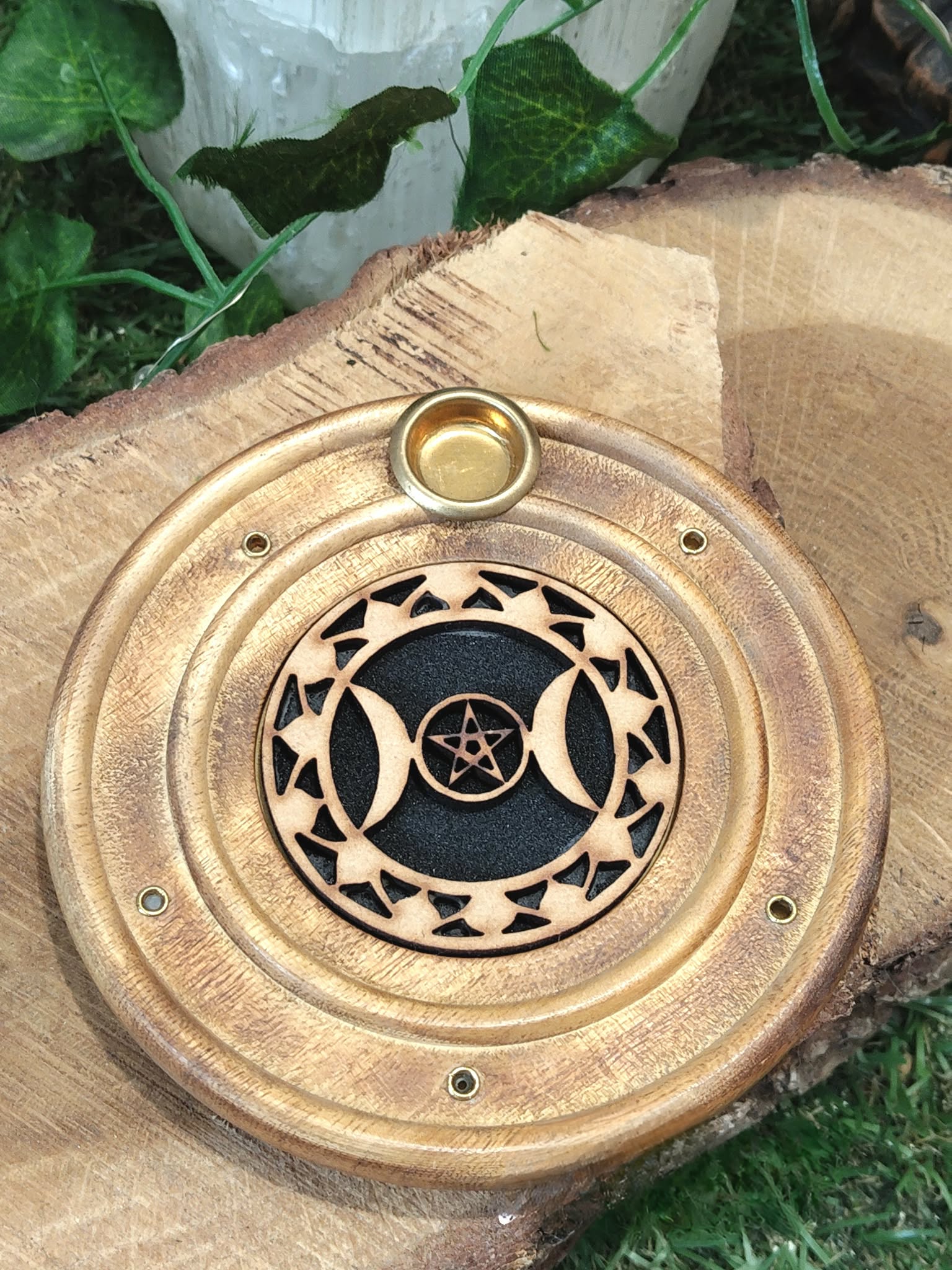 Porte-encens rond en bois motif Triple Lune, posé sur une tranche de bois avec feuillage vert en arrière-plan.