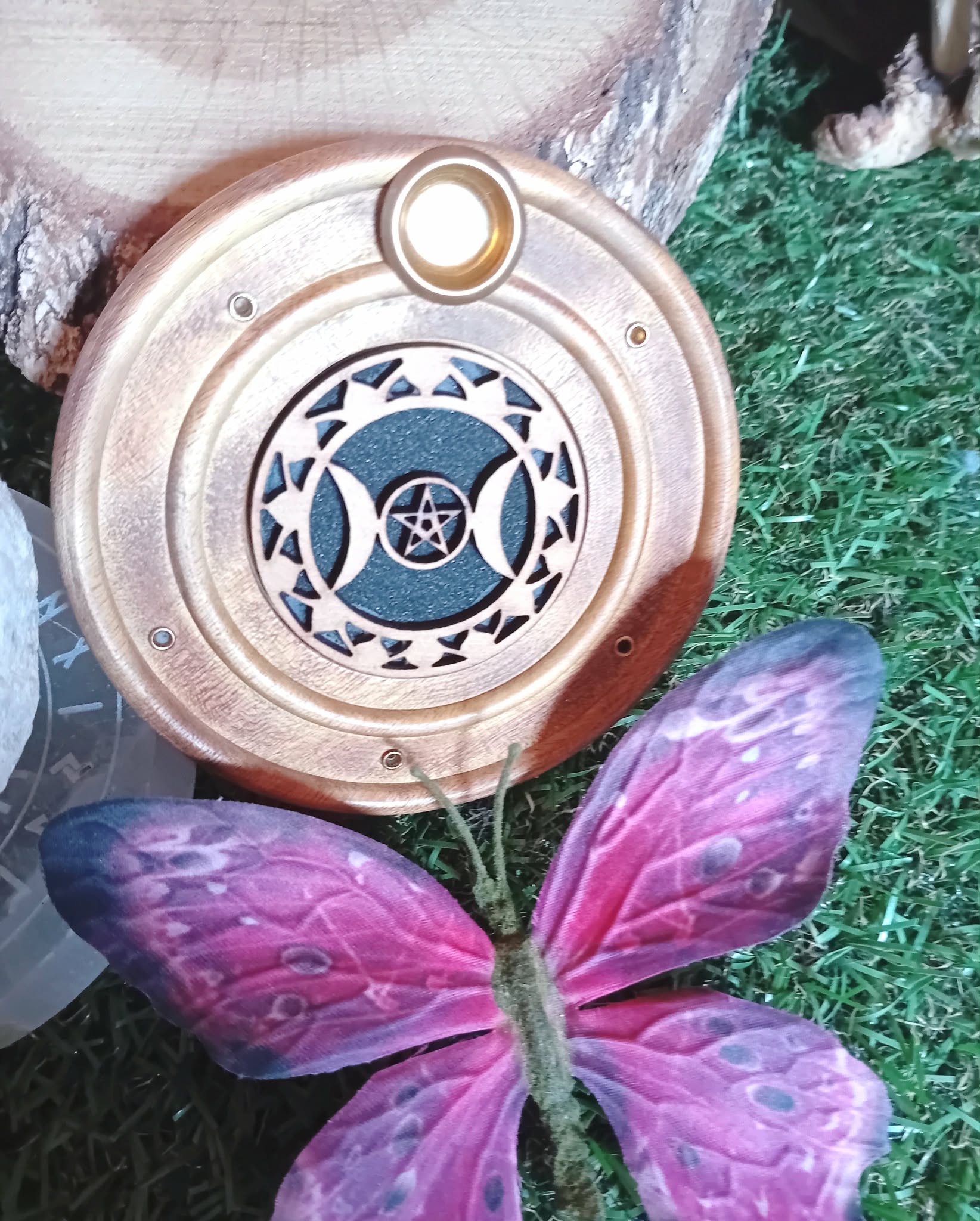 Porte-encens rond en bois motif Triple Lune, posé sur de l’herbe verte avec une décoration papillon violet au premier plan.