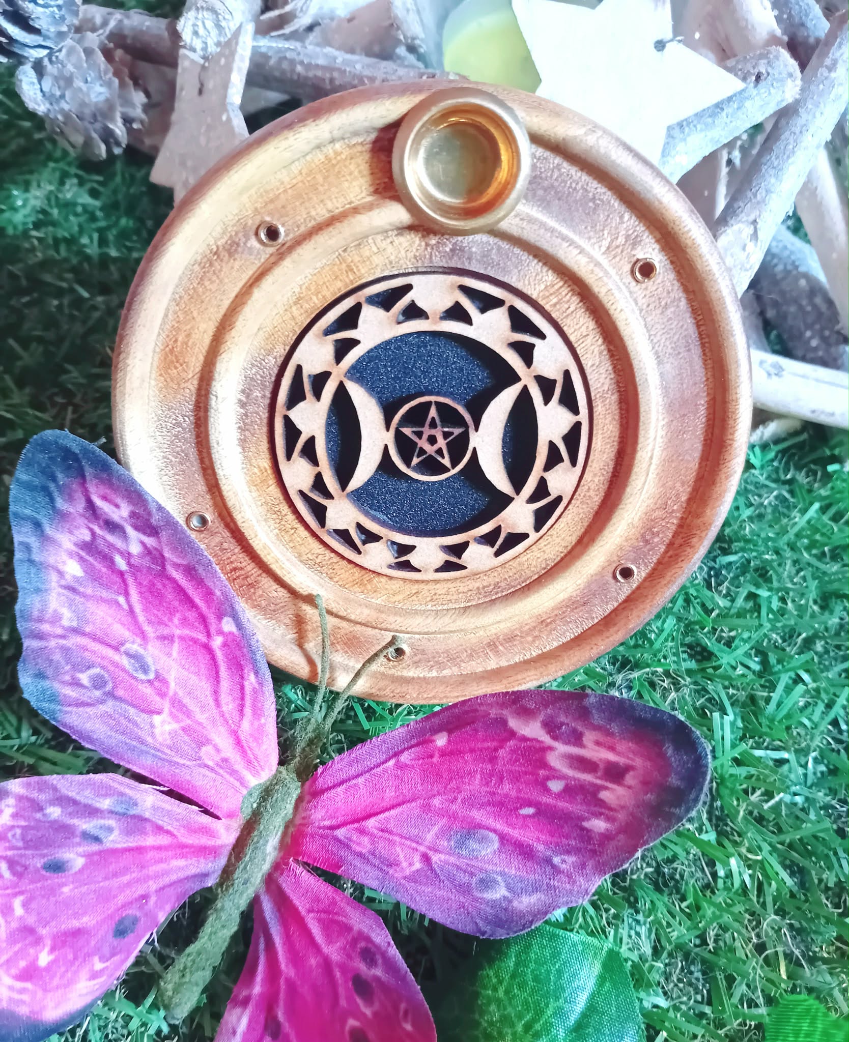 Porte-encens rond en bois motif Triple Lune, posé sur de la mousse verte avec un papillon violet en décor au premier plan