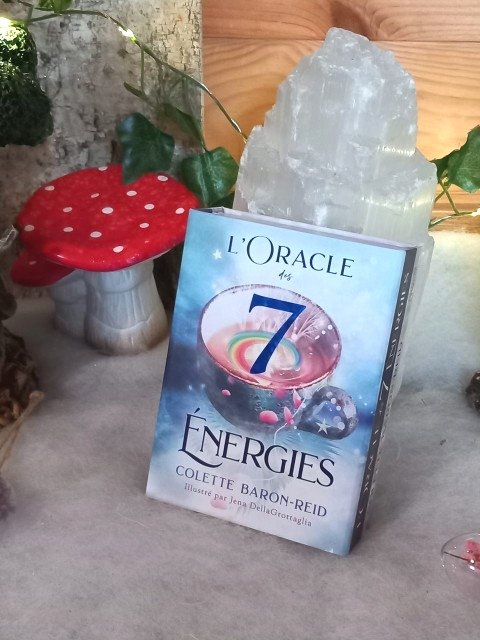 L’Oracle des 7 Énergies – coffret divinatoire en français, boîte posée sur surface grise avec décor nature (champignon décoratif rouge, feuillage) et pierre blanche en arrière-plan.