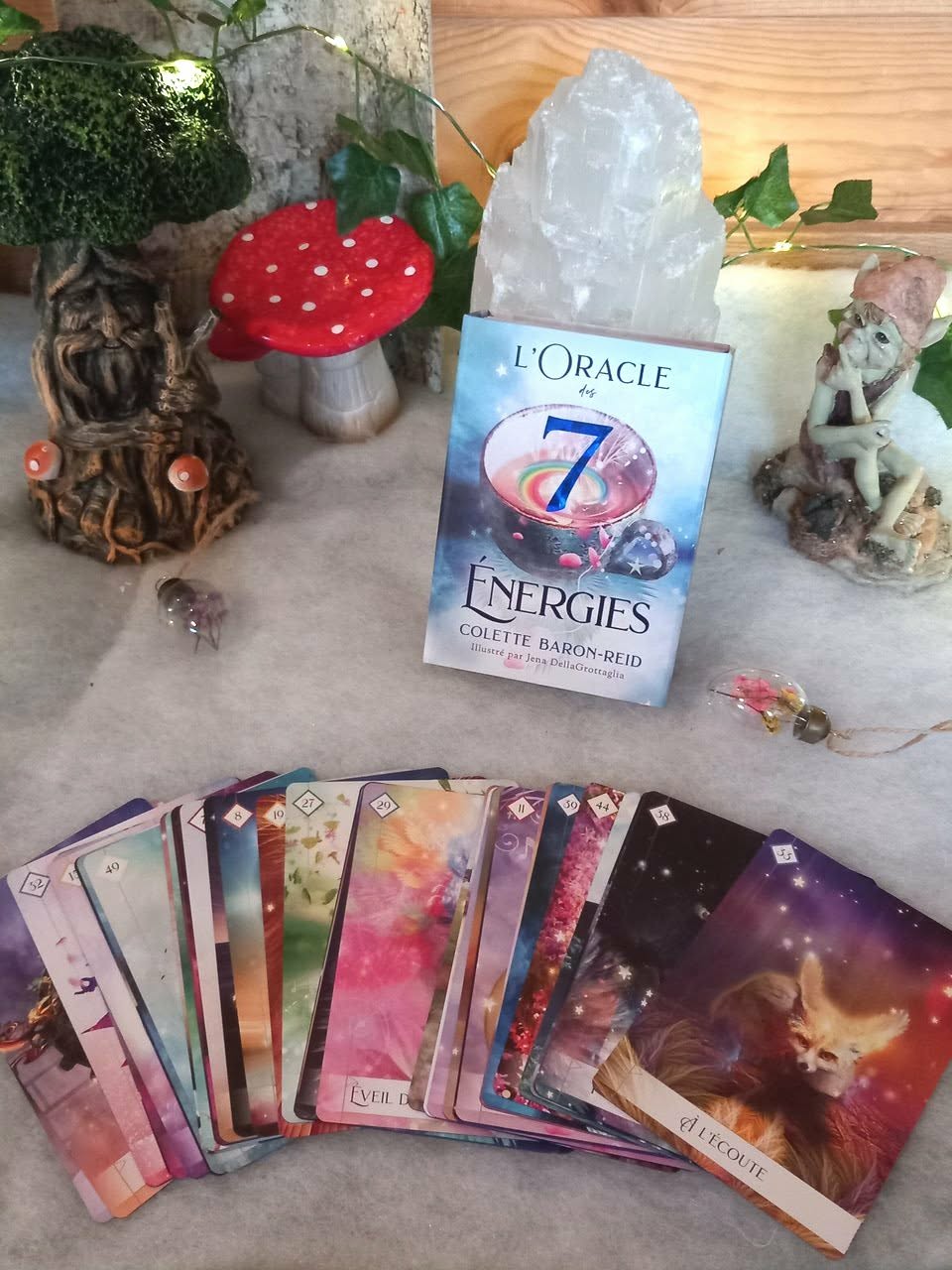L’Oracle des 7 Énergies – coffret divinatoire en français avec boîte et cartes étalées en éventail, décor nature sur table (champignon décoratif rouge, feuillage, éléments minéraux/pierre blanche) ambiance ésotérique.