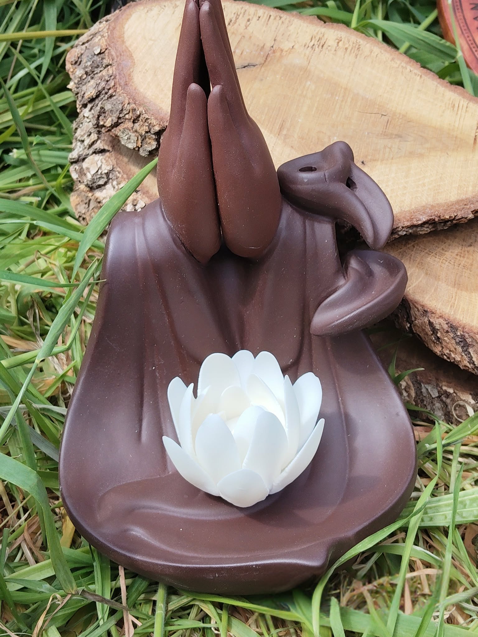 Porte-encens backflow Mains Lotus en terre cuite brun foncé, vue de dessus, posé sur herbe verte avec rondin de bois en arrière-plan.