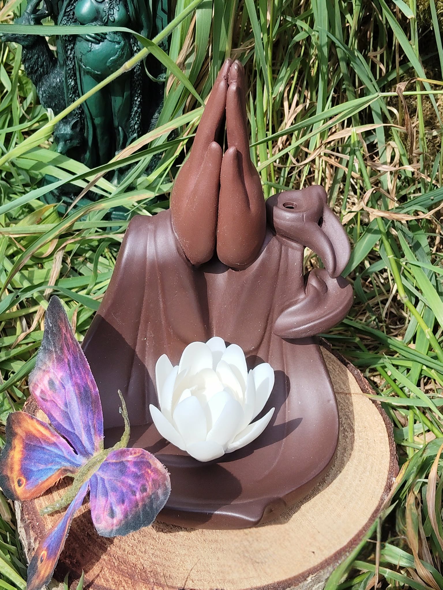 Porte-encens backflow Mains Lotus en terre cuite brun foncé, posé sur rondin de bois en extérieur, décor herbe verte avec papillon violet en avant-plan