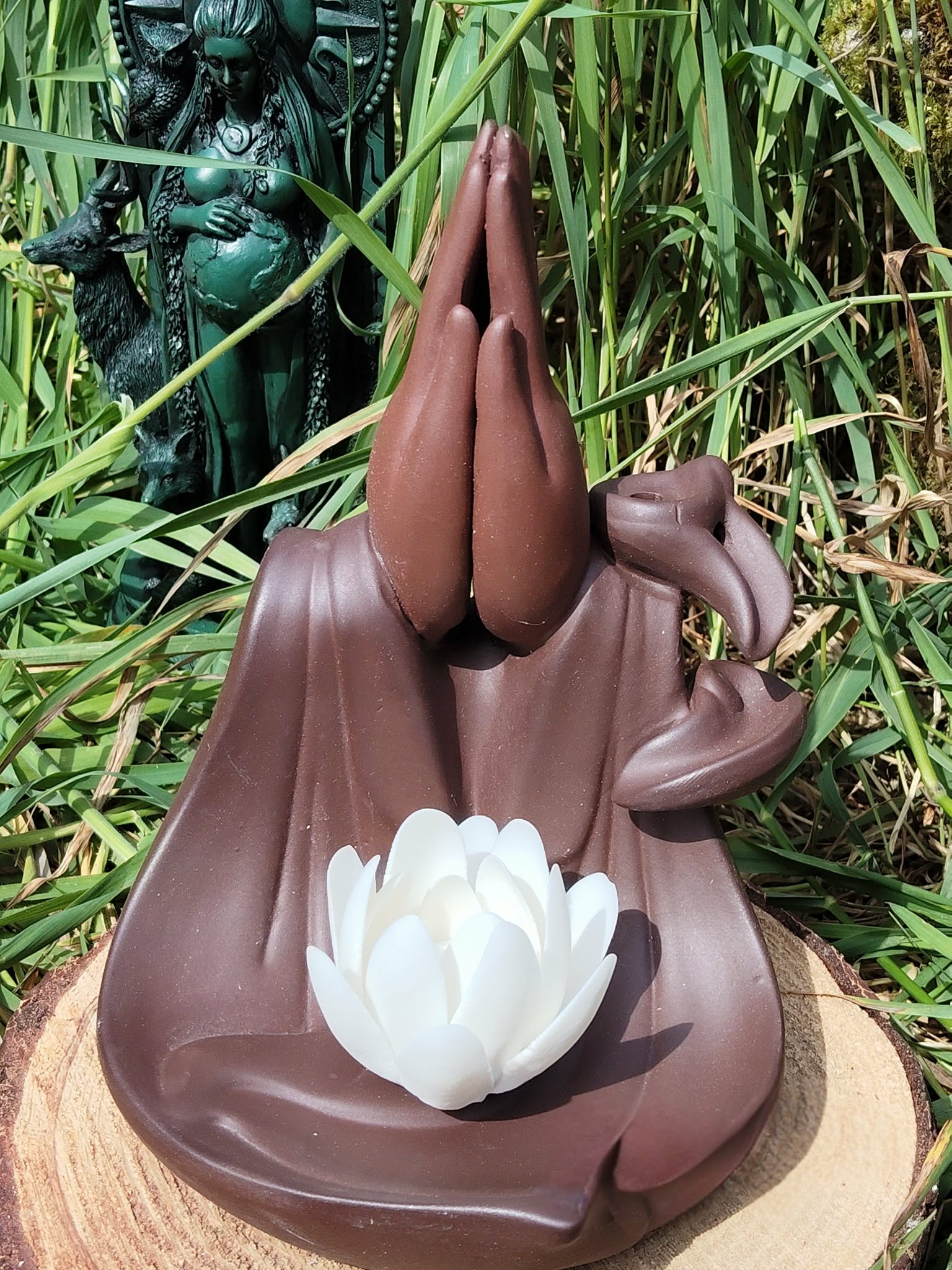 Porte-encens backflow Mains Lotus en terre cuite brun foncé, vue de face, posé sur rondin de bois, décor herbe haute en extérieur.