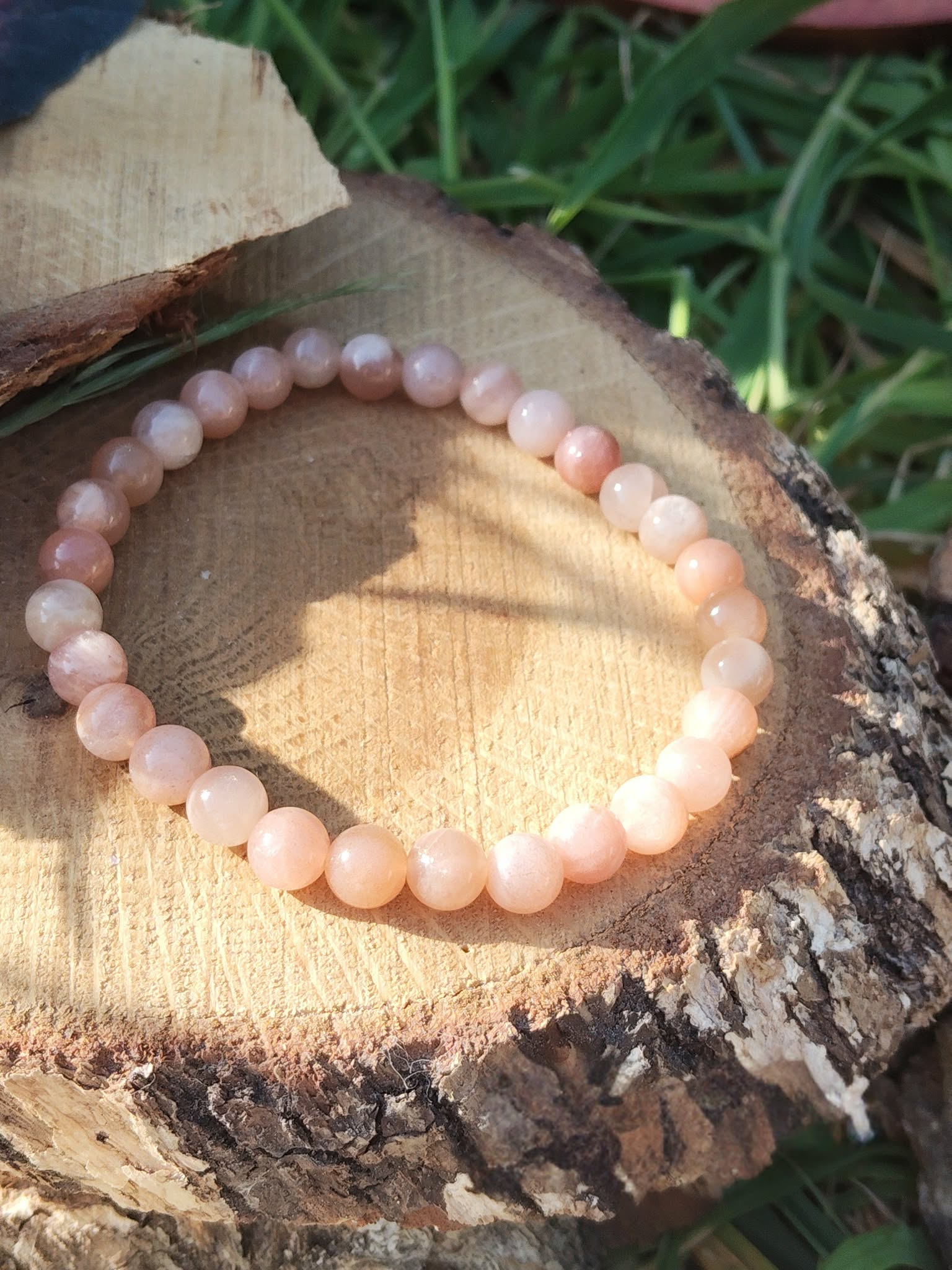 Bracelet pierre de soleil 6 mm orange pêche lumineux en perles rondes posé sur un rondin de bois, avec herbes vertes en fond, évoquant la joie, la chaleur intérieure et l’optimisme