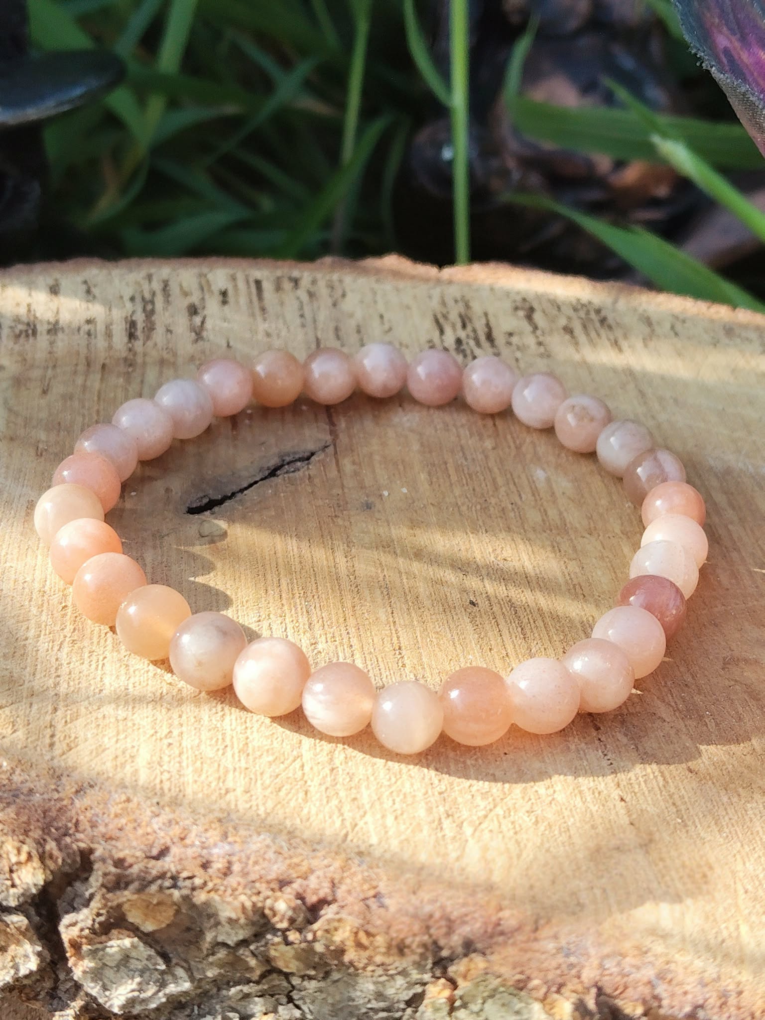 Bracelet pierre de soleil 6 mm rose orangé lumineux en perles rondes posé sur un support en bois, avec feuillage sombre en arrière-plan, évoquant l’enthousiasme, la vitalité et la confiance intérieure