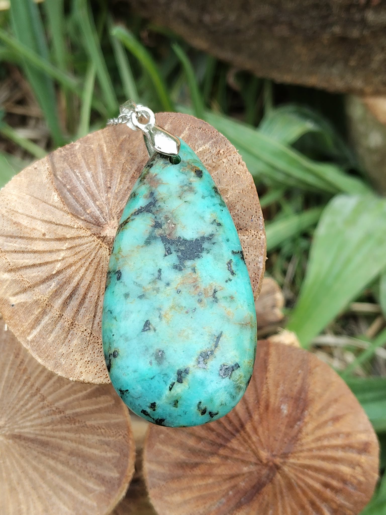 Pendentif turquoise africaine goutte bleu-vert sur bélière argentée, posé sur champignons décoratifs en bois avec feuillage, jaspe africain évoquant la stabilité émotionnelle, l’aisance et le calme du cœur.