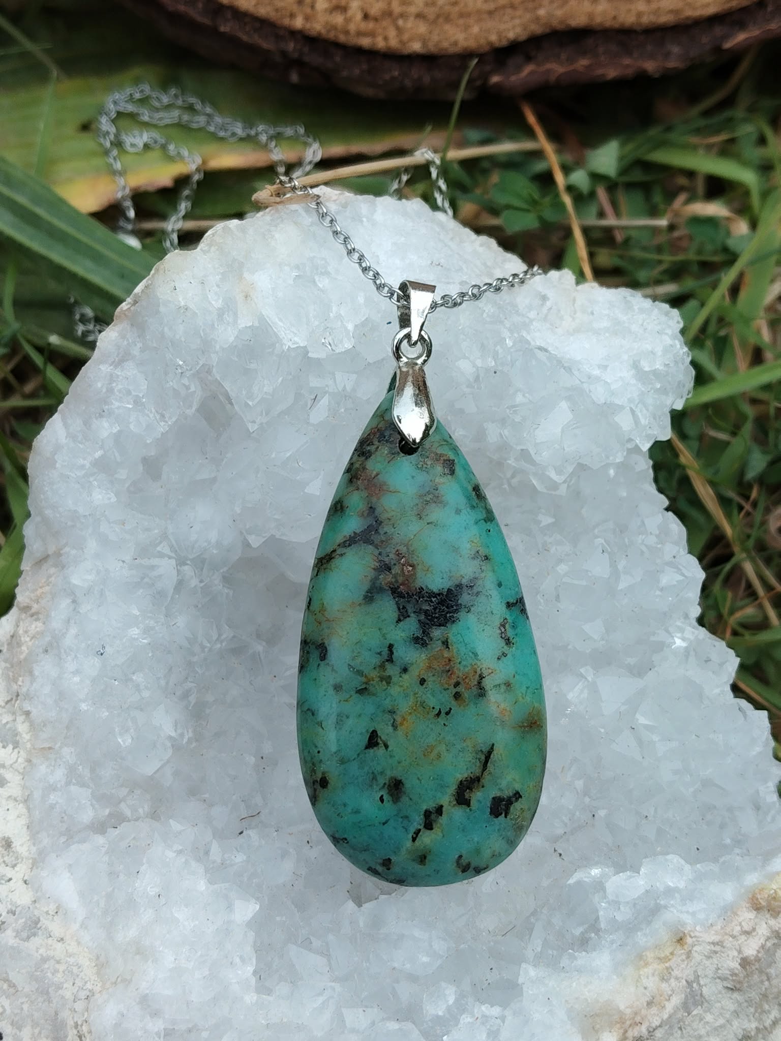 Pendentif turquoise africaine forme goutte sur bélière argentée, suspendu sur amas de quartz brut avec décor herbe et bois, jaspe africain associé au recentrage, à l’expression calme et à l’équilibre intérieur.