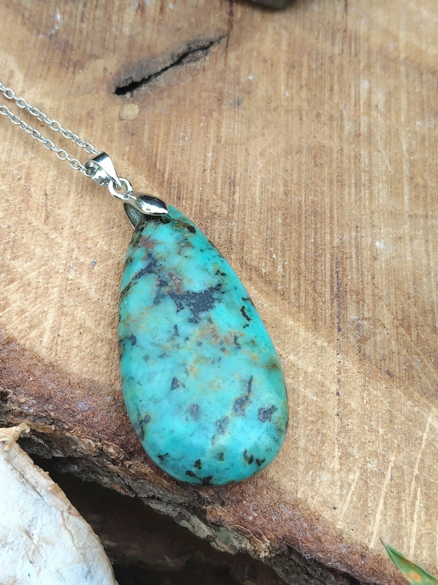 Pendentif turquoise africaine goutte bleu-vert sur bélière argentée, posé sur bois clair, jaspe africain lié à la présence à soi, à la souplesse émotionnelle et à la communication calme.