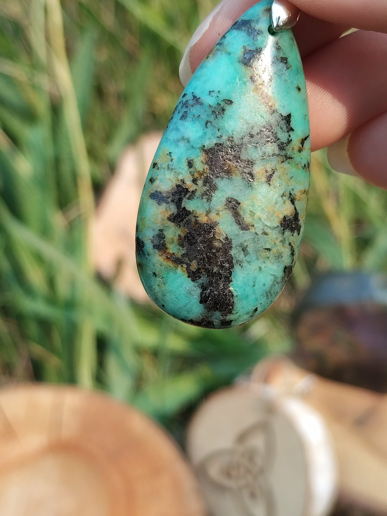 Gros plan sur pendentif turquoise africaine goutte bleu-vert marbrée de noir, tenu entre les doigts devant feuillage, jaspe africain évoquant la lucidité, le recentrage et la confiance tranquille.