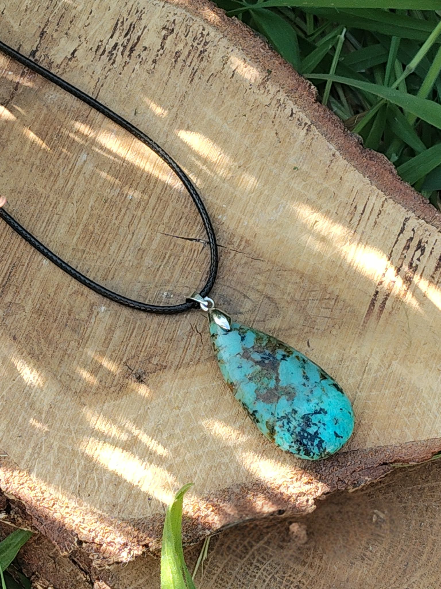Collier cordon noir avec pendentif turquoise africaine goutte bleu-vert marbrée posé sur rondelle de bois avec herbe, jaspe africain évoquant l’ancrage, la clarté intérieure et l’expression posée.