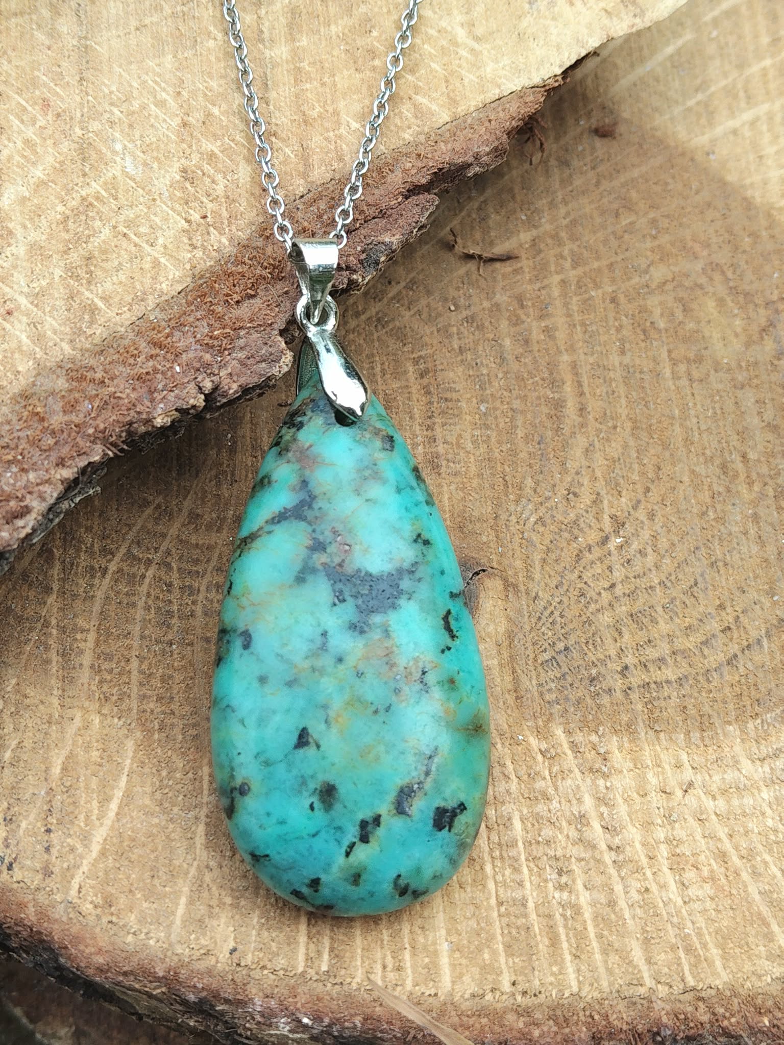 Pendentif turquoise africaine forme goutte bleu-vert sur bélière argentée, posé sur bois clair, jaspe africain associé à la douceur intérieure, à la communication apaisée et à l’harmonie émotionnelle.
