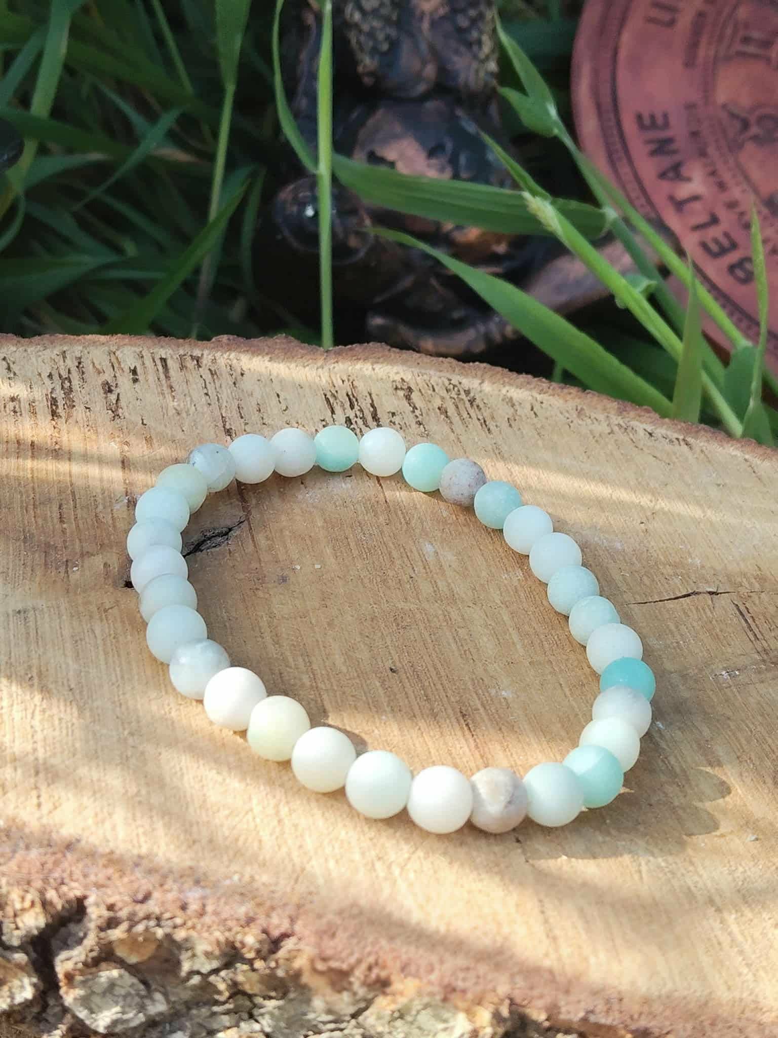 Bracelet amazonite multicolore en perles 6 mm, posé sur rondin de bois avec feuillage vert et détail naturel en fond ; pierre associée à l’apaisement, à la confiance et à l’équilibre émotionnel.