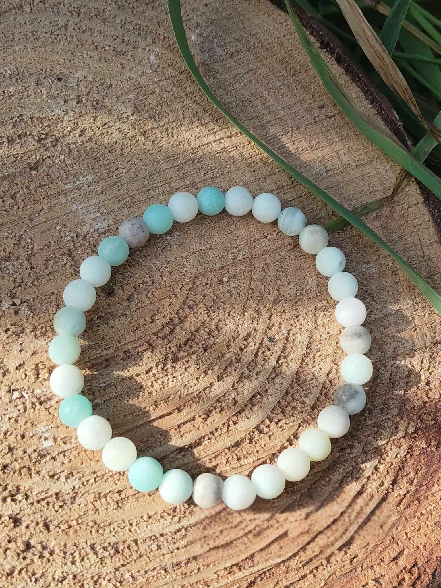 Bracelet amazonite multicolore en perles 6 mm, photographié sur bois texturé avec feuillage vert en arrière-plan ; pierre associée à l’apaisement, à l’expression et à l’équilibre émotionnel.