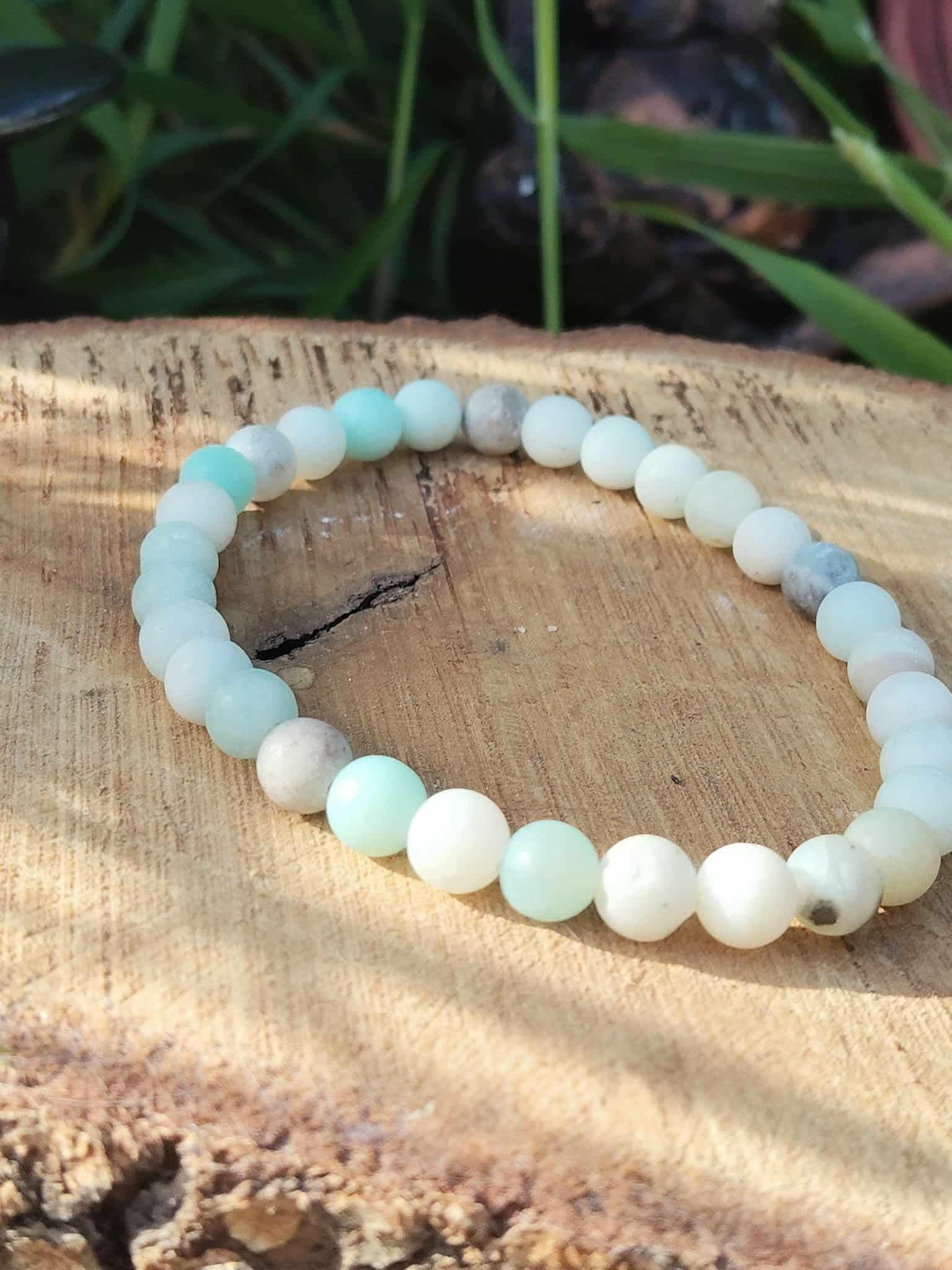 Bracelet amazonite multicolore en perles 6 mm, posé sur rondin de bois avec feuillage vert en arrière-plan ; pierre associée à l’apaisement, à l’équilibre émotionnel et à une communication plus douce.