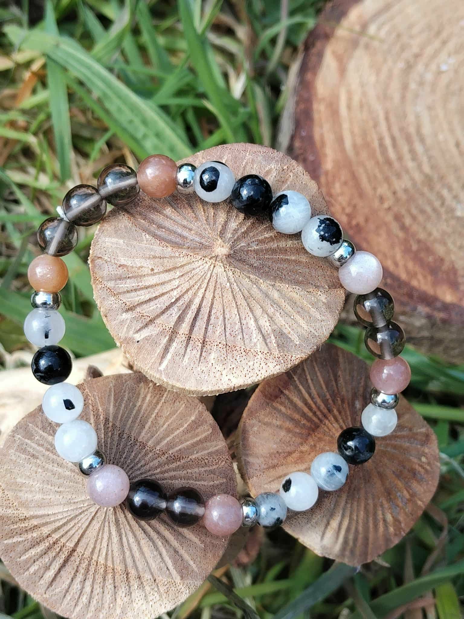 Bracelet Deuil mis en scène sur rondins de bois avec feuillage vert, aide à traverser les vagues émotionnelles et à retrouver un appui plus stable