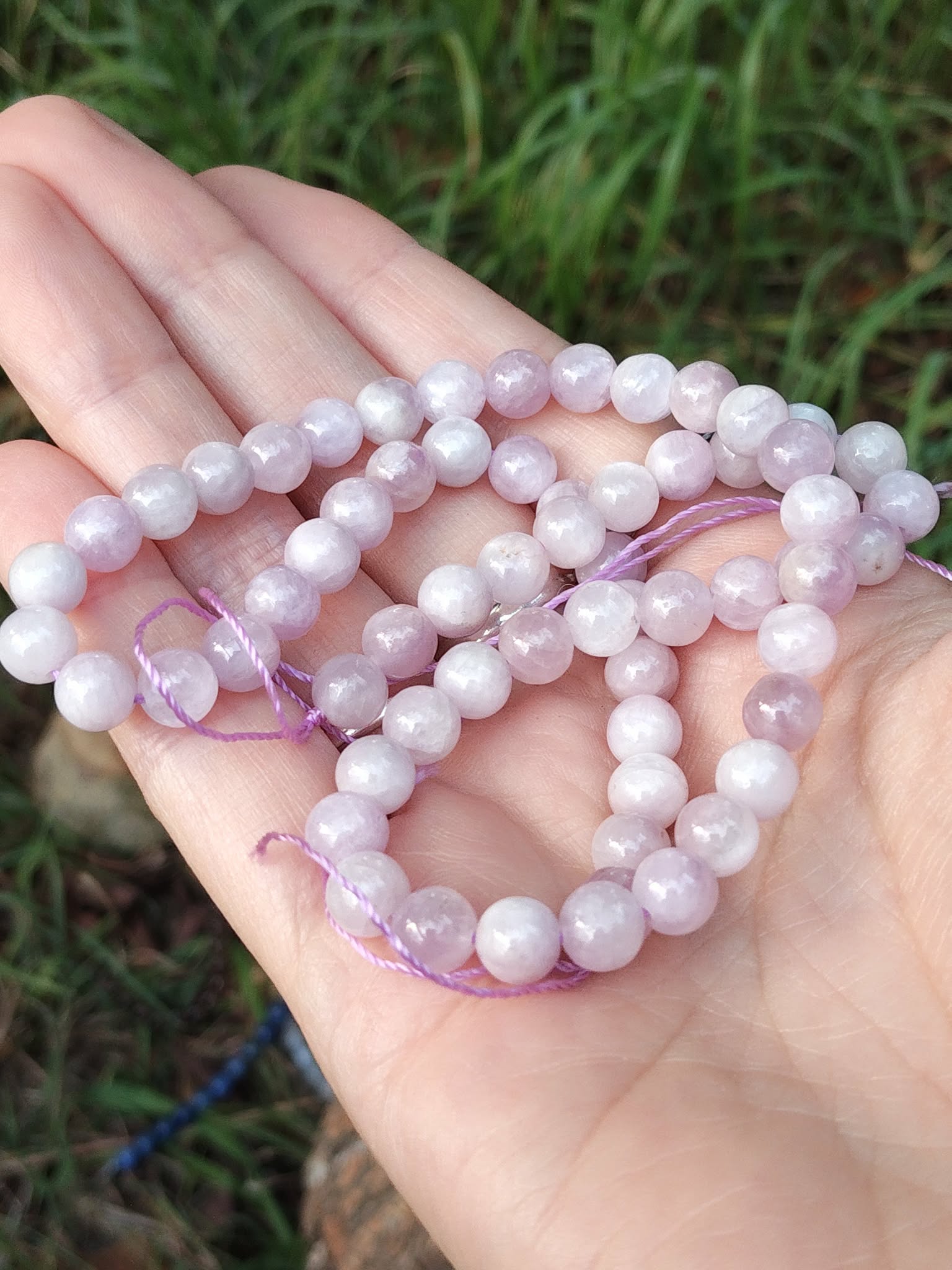 Fil de perles kunzite 6 mm tenu en main devant feuillage, tons rose pastel et lilas clair pour délicatesse intérieure, appui émotionnel et douceur stable