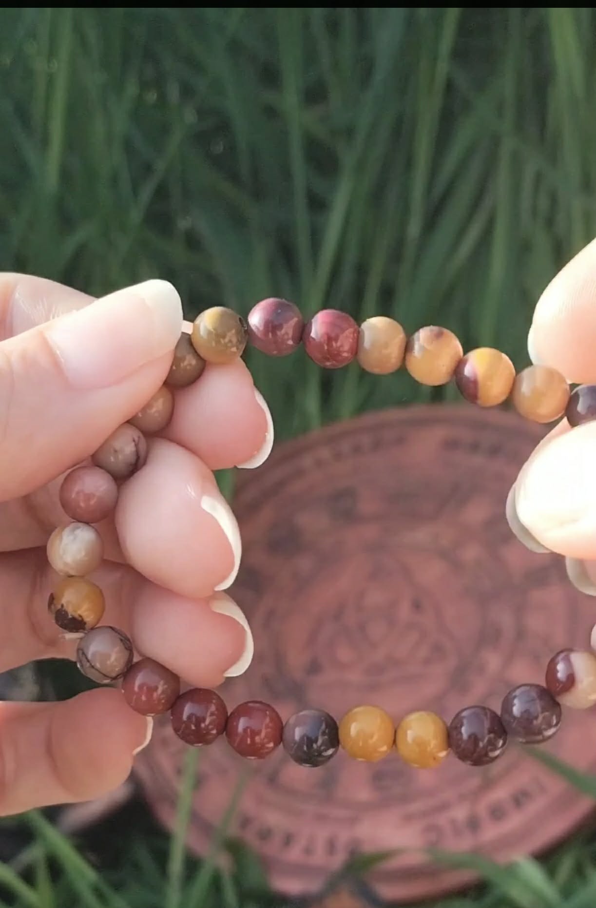 Bracelet mookaïte 6 mm porté au poignet, perles ocre, beige, bordeaux et prune, avec plaque en cuivre triquetra et feuillage vert, assurance, centrage et équilibre intérieur