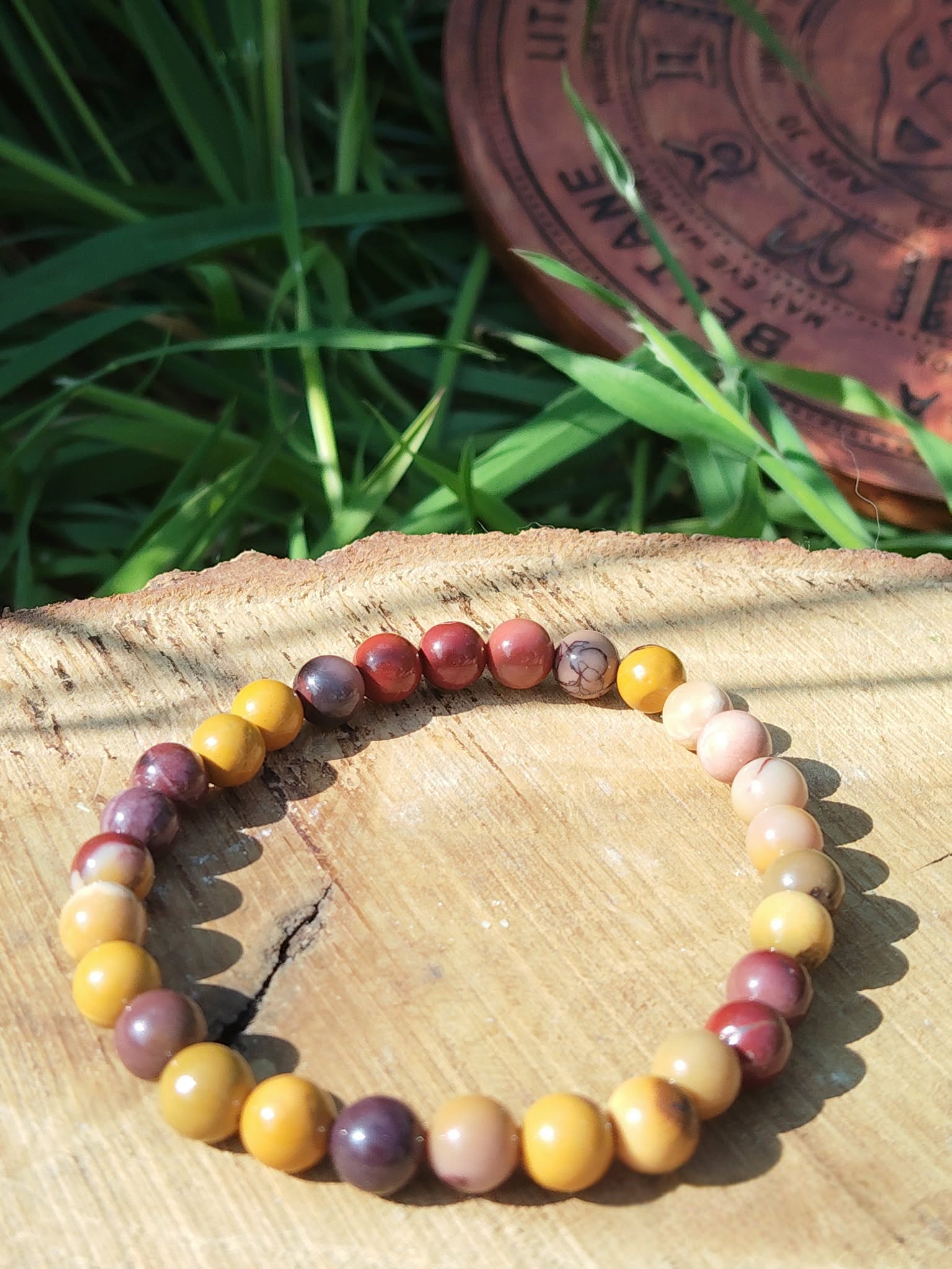 Bracelet mookaïte 6 mm sur planche en bois avec feuillage vert et disque gravé, perles ocre, crème, bordeaux et prune pour stabilité, élan vital et assurance sereine