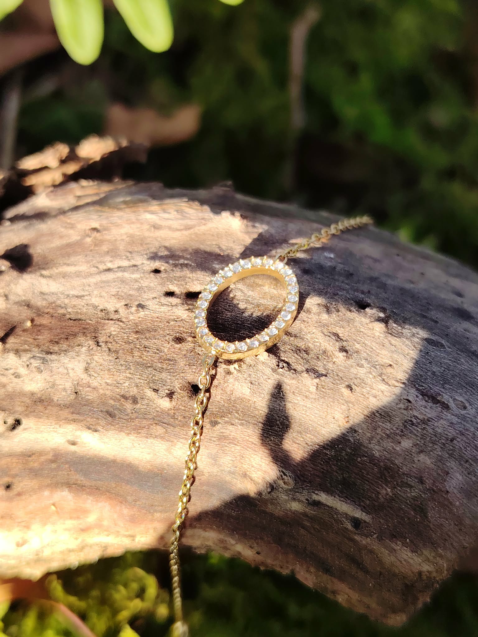 Bracelet chaîne ovale strass doré sur tronc de bois clair, mousse et feuillage en arrière-plan, bracelet fin femme tendance