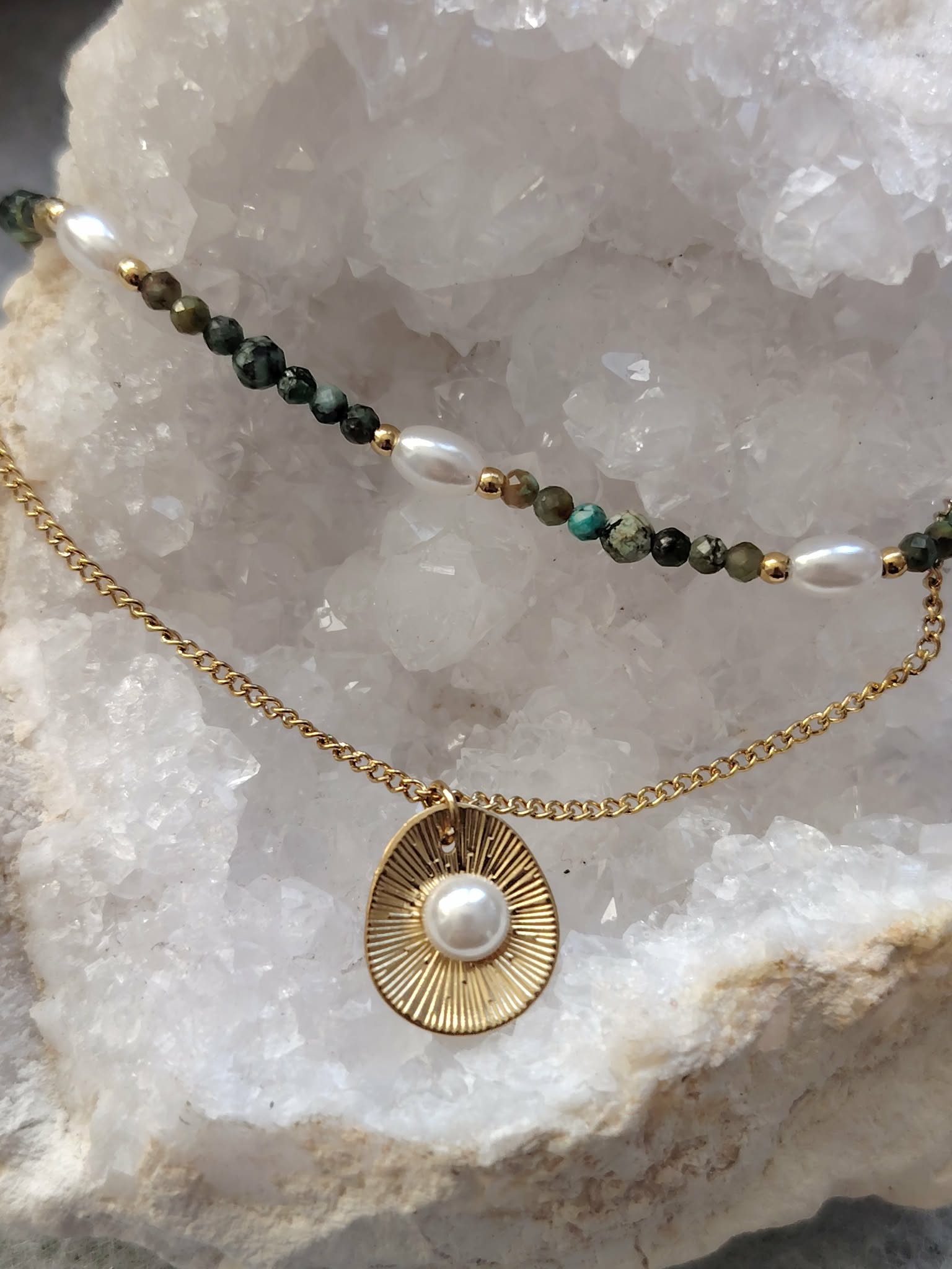 Collier double chaîne dorée en acier inoxydable avec perles blanches, pierres vertes et médaillon nacré, présenté sur buste blanc, gros plan sur le pendentif, fond clair et amas de quartz en décor.