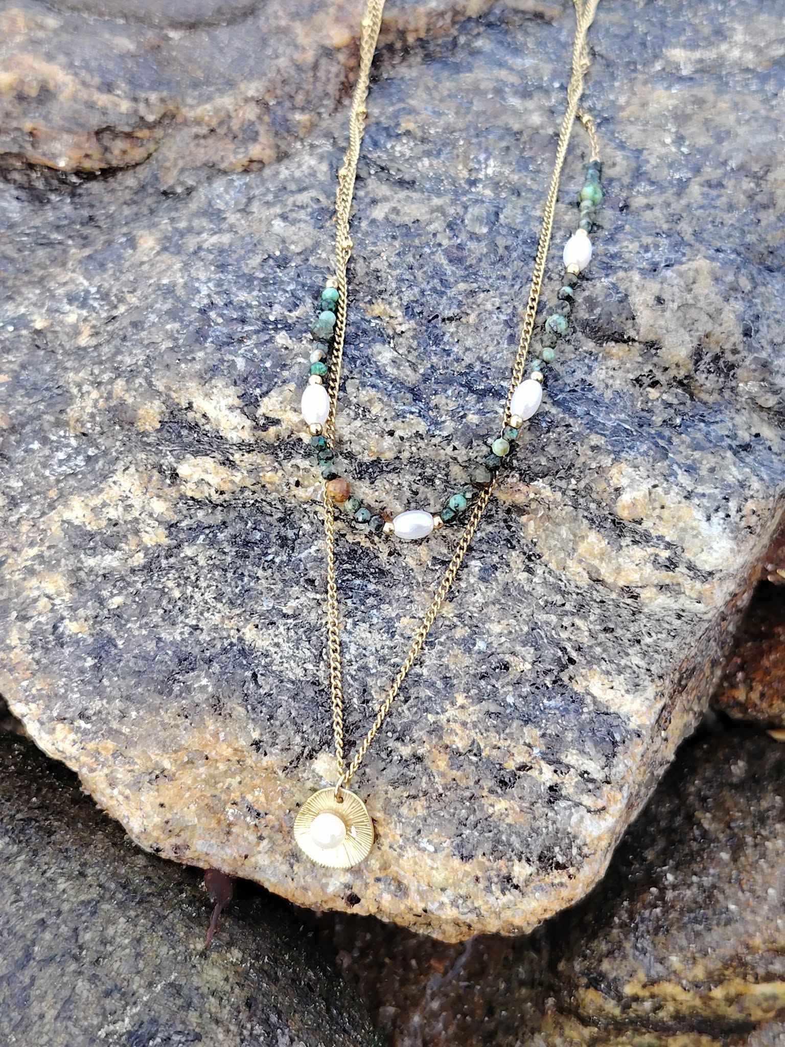 Collier double chaîne dorée en acier inoxydable avec perles blanches, pierres vertes et médaillon nacré, suspendu sur une pierre gris bleuté texturée, décor naturel, lumière douce.