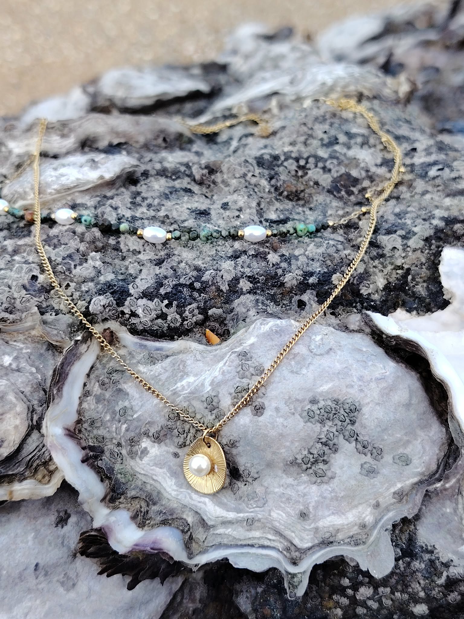 Collier double chaîne dorée en acier inoxydable avec perles blanches, pierres vertes et médaillon nacré, photographié en Bretagne, posé sur un rocher avec une coquille type huître en décor, ambiance minérale, lumière naturelle douce.